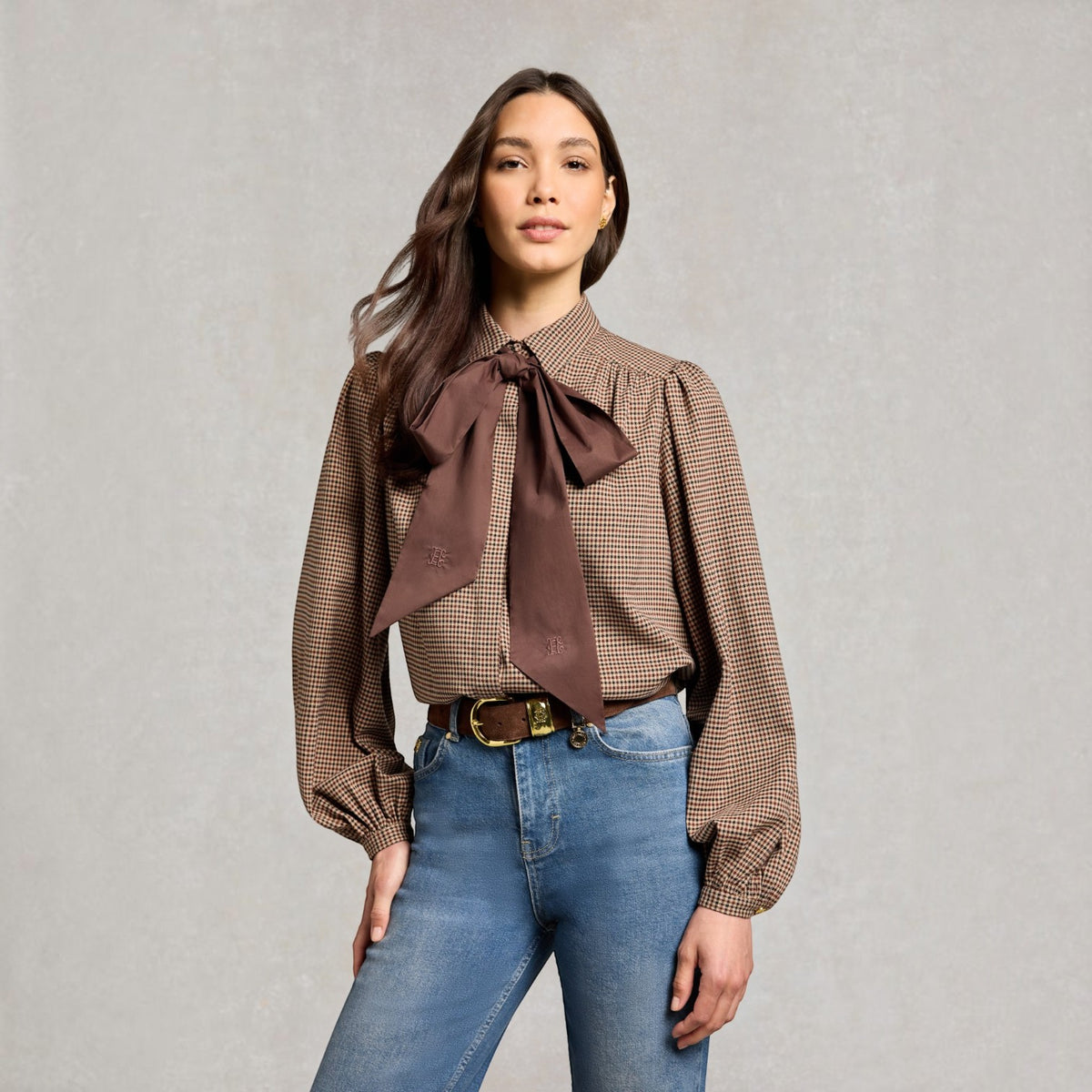 A woman modeling a brown and beige checkered long-sleeve blouse with a large matching brown bow tie at the collar, paired with blue jeans and a gold belt, against a textured gray studio background.