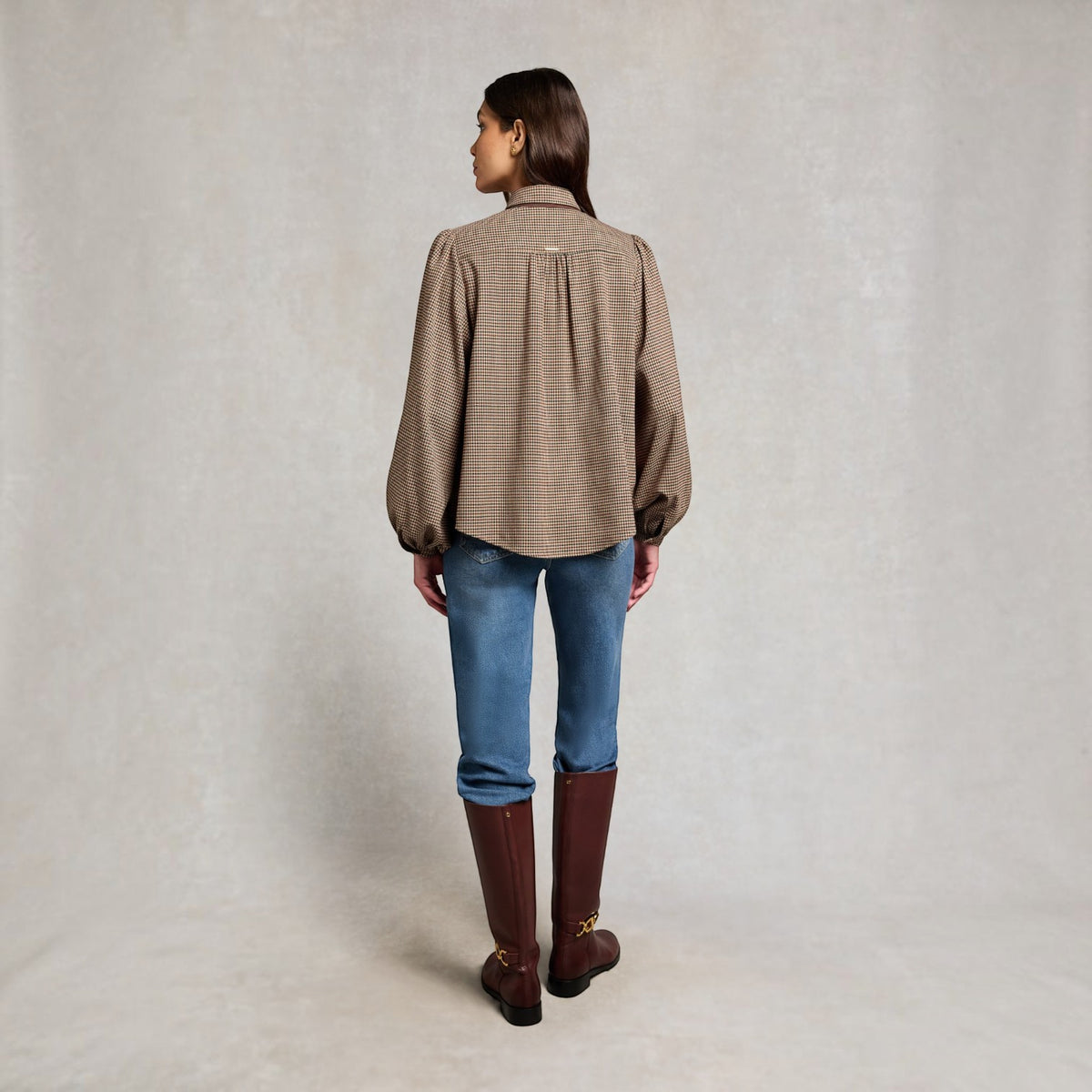 A woman is shown from the back wearing a brown and beige checkered long-sleeve blouse, blue jeans, and brown knee-high boots against a neutral studio backdrop.