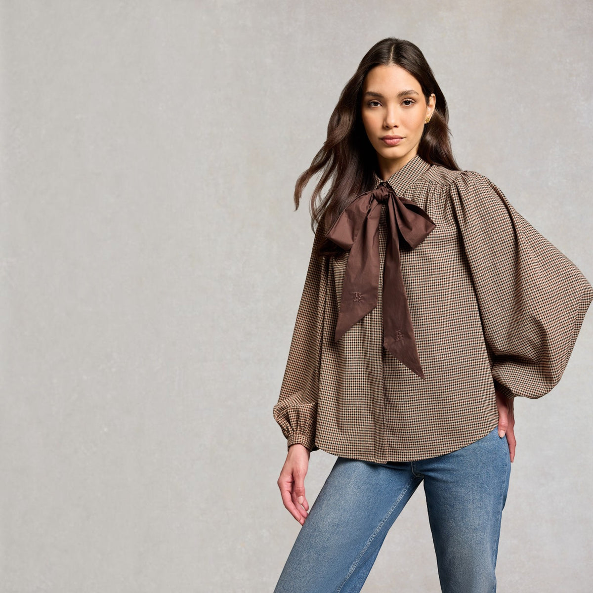A woman modeling a brown and beige houndstooth blouse with a large dark brown bow at the collar, paired with blue jeans, against a textured light gray background.
