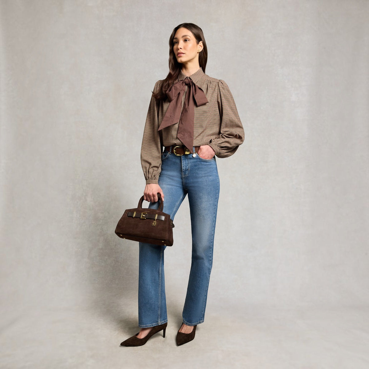A woman stands against a textured light gray studio backdrop wearing a brown long-sleeve blouse with a large bow at the collar, blue jeans, dark brown heels, and holding a brown handbag.