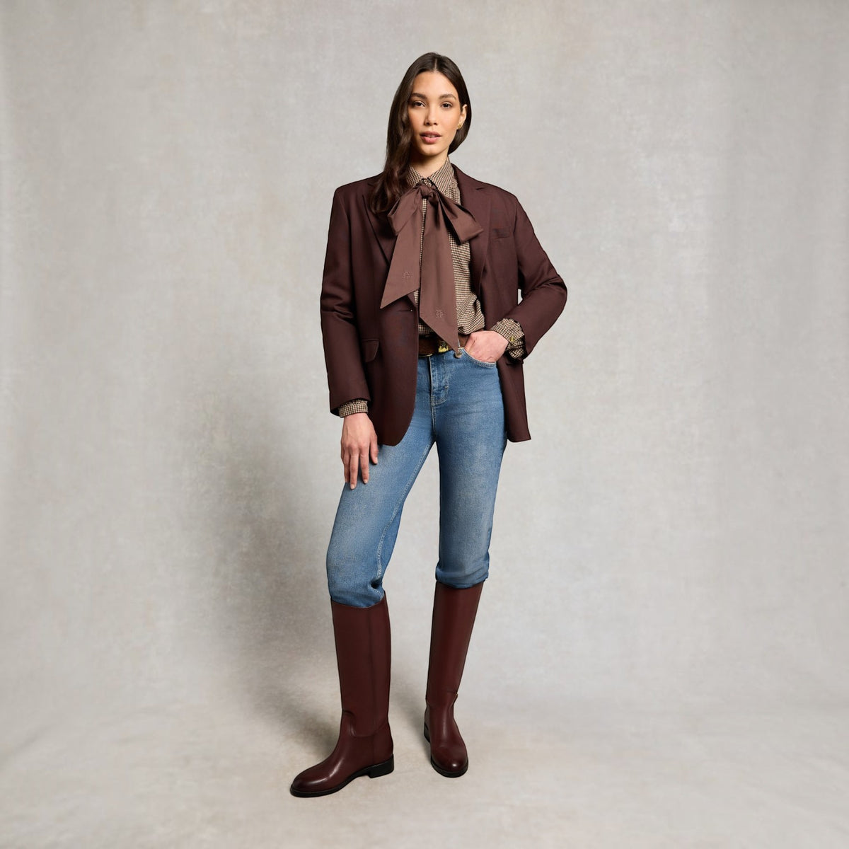 A model stands against a textured light gray studio backdrop wearing a dark brown blazer, a matching brown bow tie blouse, blue jeans, and dark brown knee-high boots.