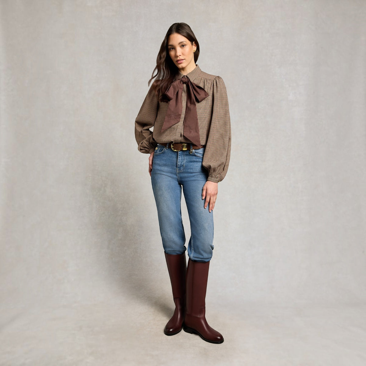 A woman stands against a textured light gray studio backdrop wearing a brown patterned blouse with a large dark brown bow at the collar, blue jeans, and dark brown knee-high boots.