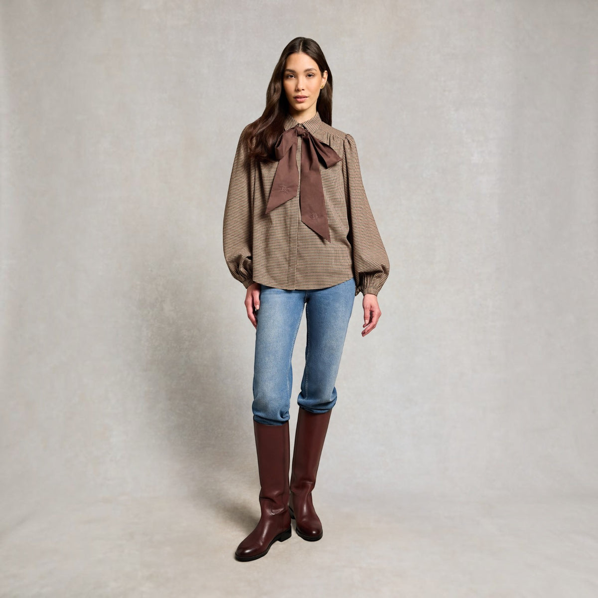 A woman stands against a textured light gray studio backdrop wearing a brown patterned blouse with a large dark brown bow at the collar, blue jeans, and dark brown knee-high boots.