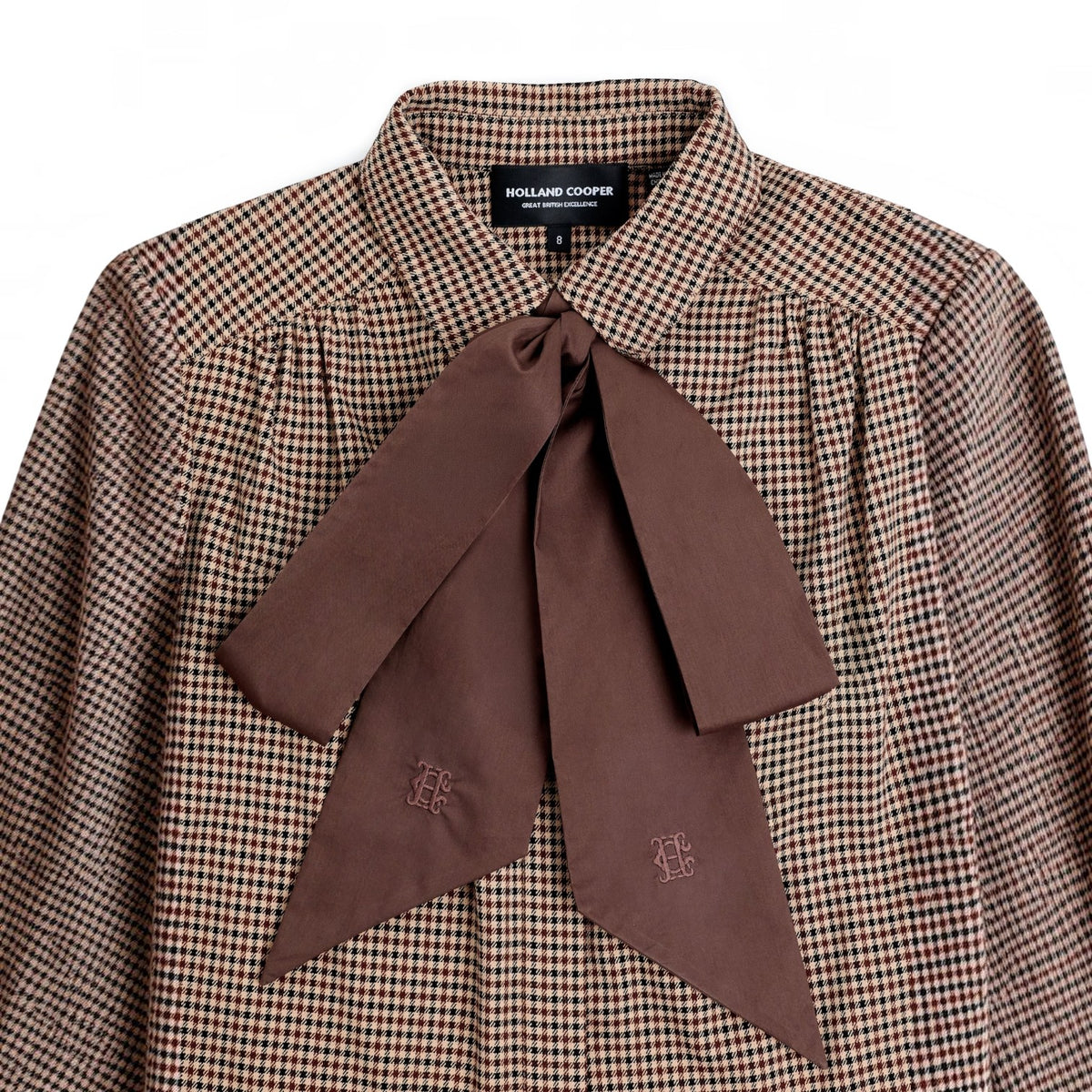 A close-up of a collared blouse with a brown and beige gingham pattern and a large brown satin bow at the front, featuring a visible Holland Cooper neck label.