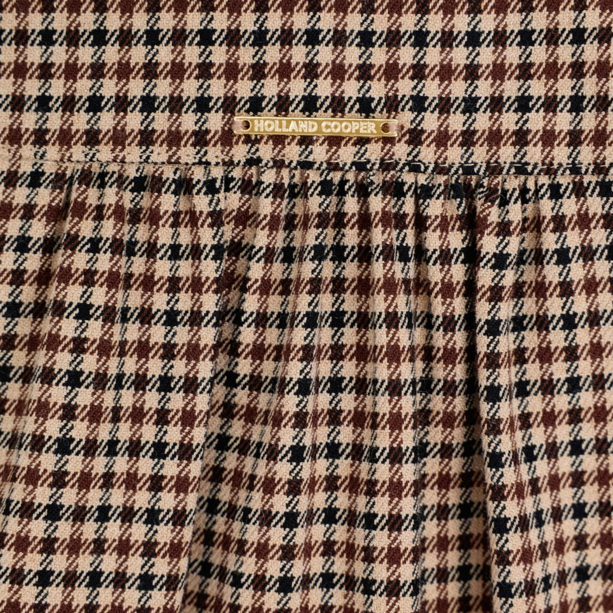 A close-up of a plaid fabric pattern in beige, brown, and black, with a small gold-tone metal brand plaque reading 'HOLLAND COOPER' near the top center.