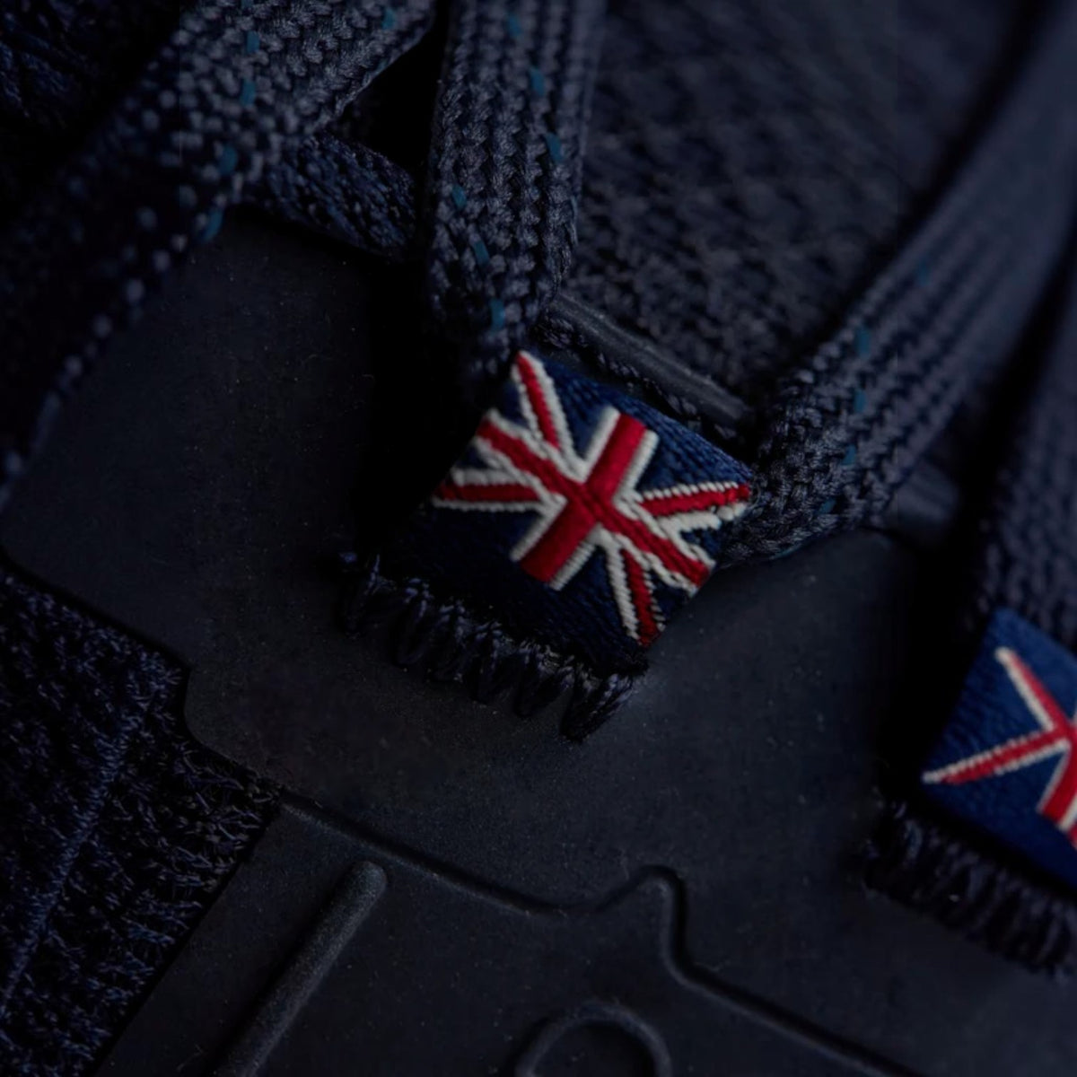 Close-up of dark navy blue woven laces with embroidered Union Jack flags in red, white, and blue attached near the tongue area of a shoe.