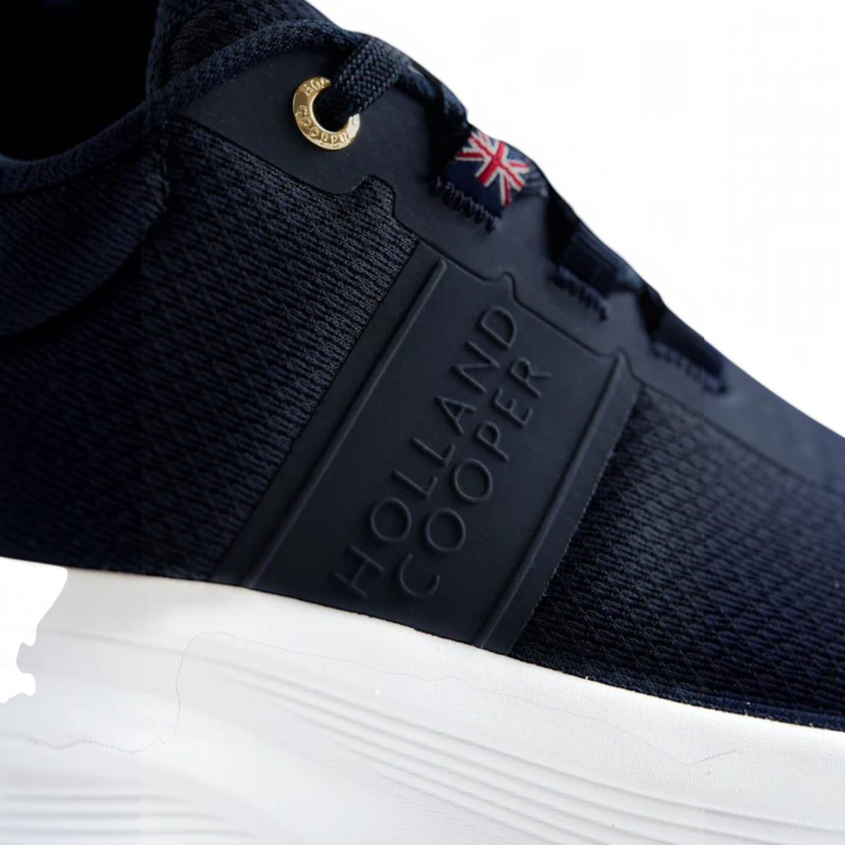 Close-up of a black mesh sneaker with a black rubber side panel embossed with 'HOLLAND COOPER', a white sole, a gold lace loop, and a small Union Jack eyelet detail on a white background.
