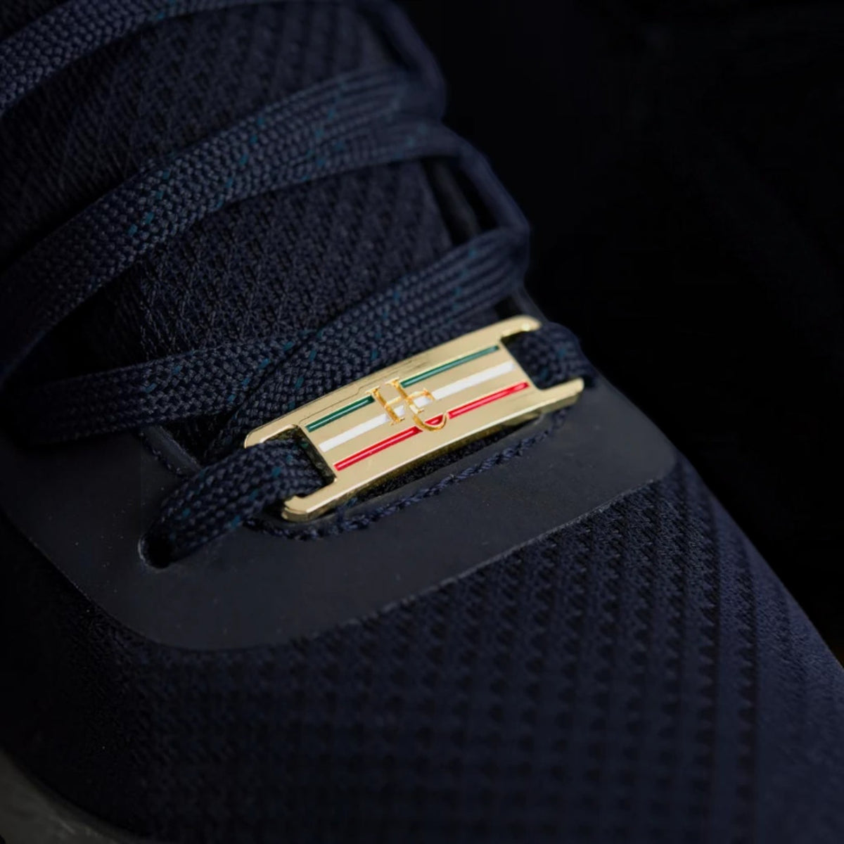 Close-up of a navy blue athletic shoe with a gold metal lace lock featuring the Italian flag colors (green, white, and red) on the tongue.