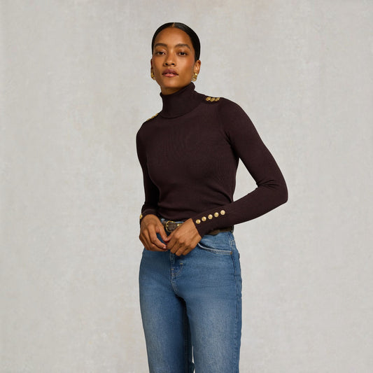 A woman modeling a dark brown ribbed turtleneck sweater with gold button details on the shoulders and cuffs, paired with blue jeans, against a textured light gray background.