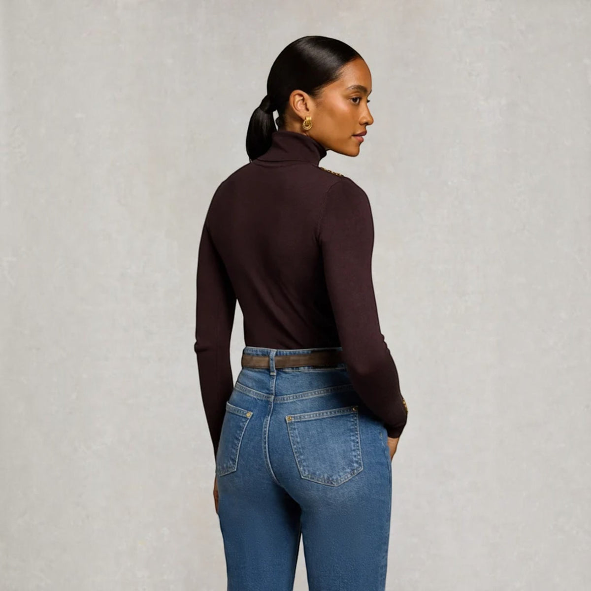 A woman wearing a dark plum long-sleeve turtleneck top and blue jeans, photographed from the back and side against a textured light gray studio background.
