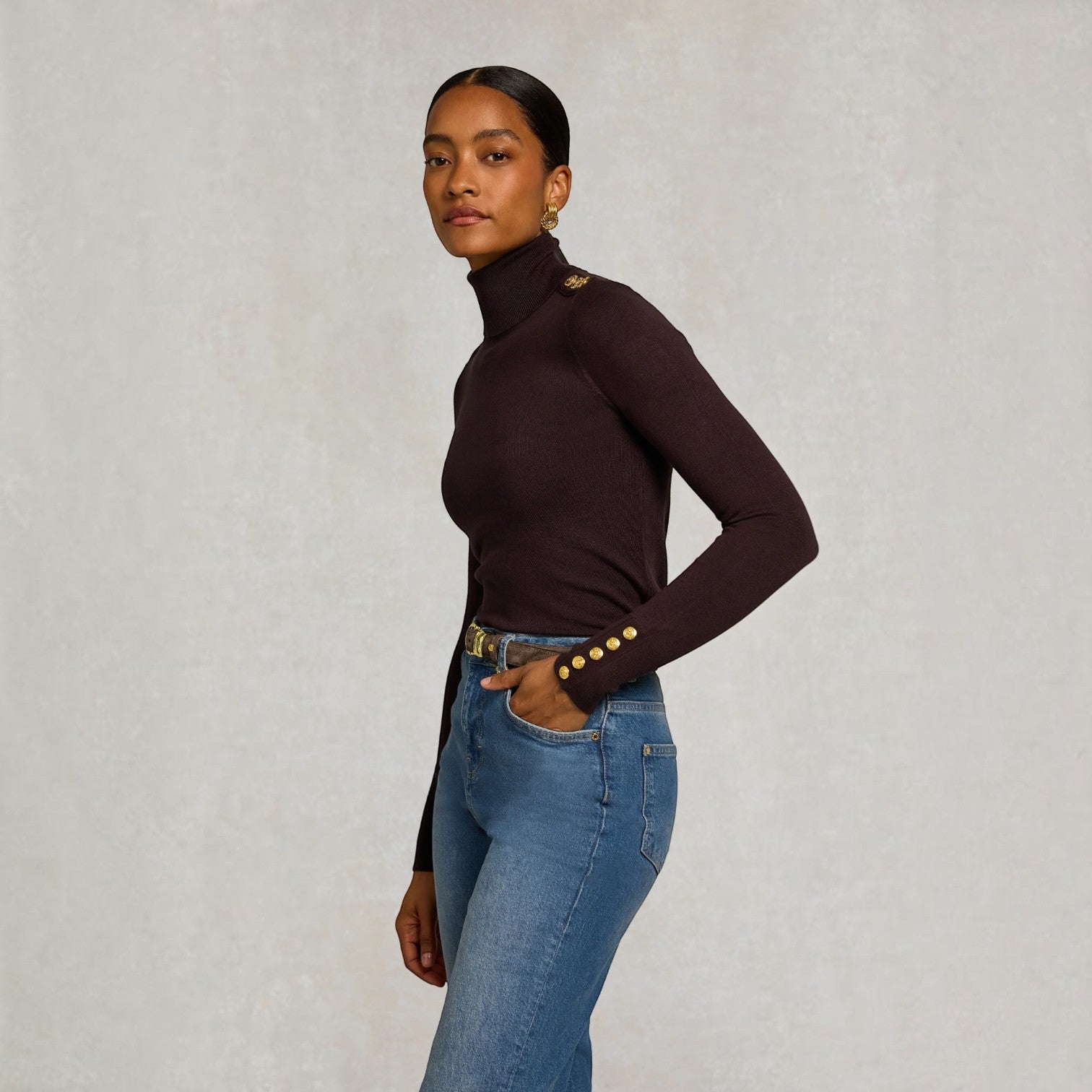 A woman modeling a dark brown ribbed turtleneck top with gold button details on the shoulders and cuffs, paired with blue jeans, against a textured light gray background.