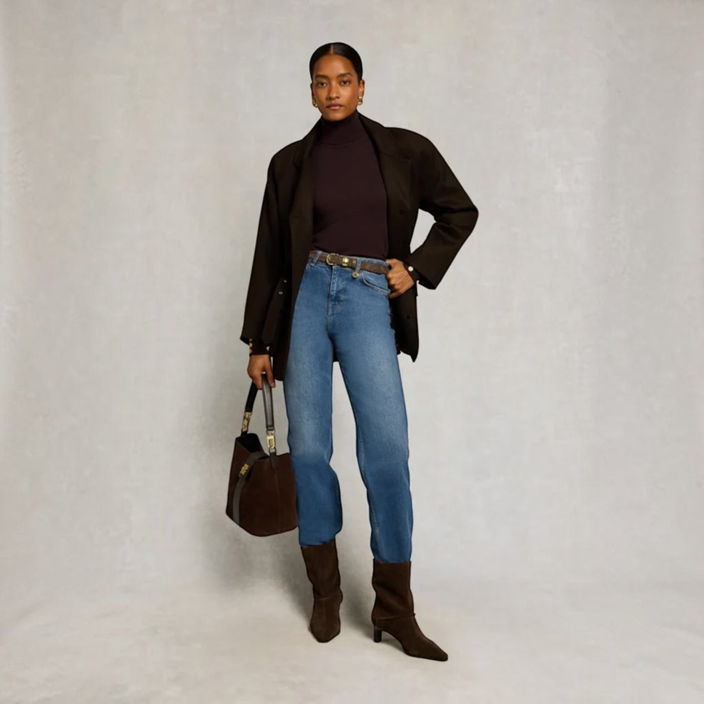 A model stands against a light gray studio backdrop wearing a dark brown blazer, dark plum turtleneck, blue jeans, and dark brown heeled boots, while holding a dark brown handbag.