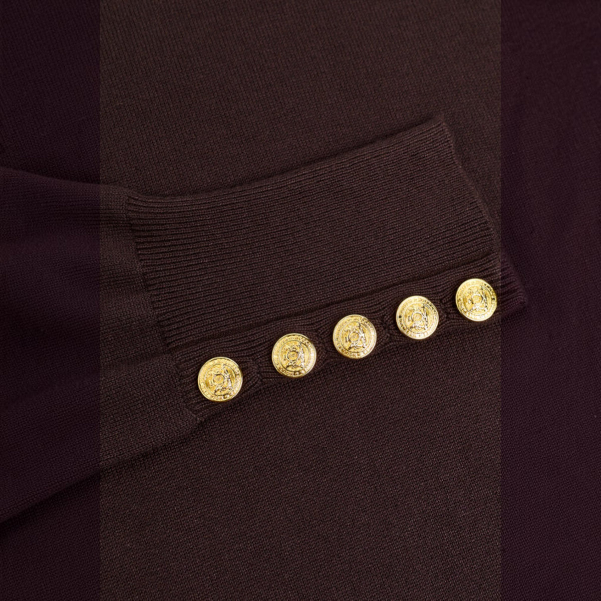 Close-up of a dark brown ribbed knit sleeve cuff featuring five decorative gold buttons with embossed circular motifs.