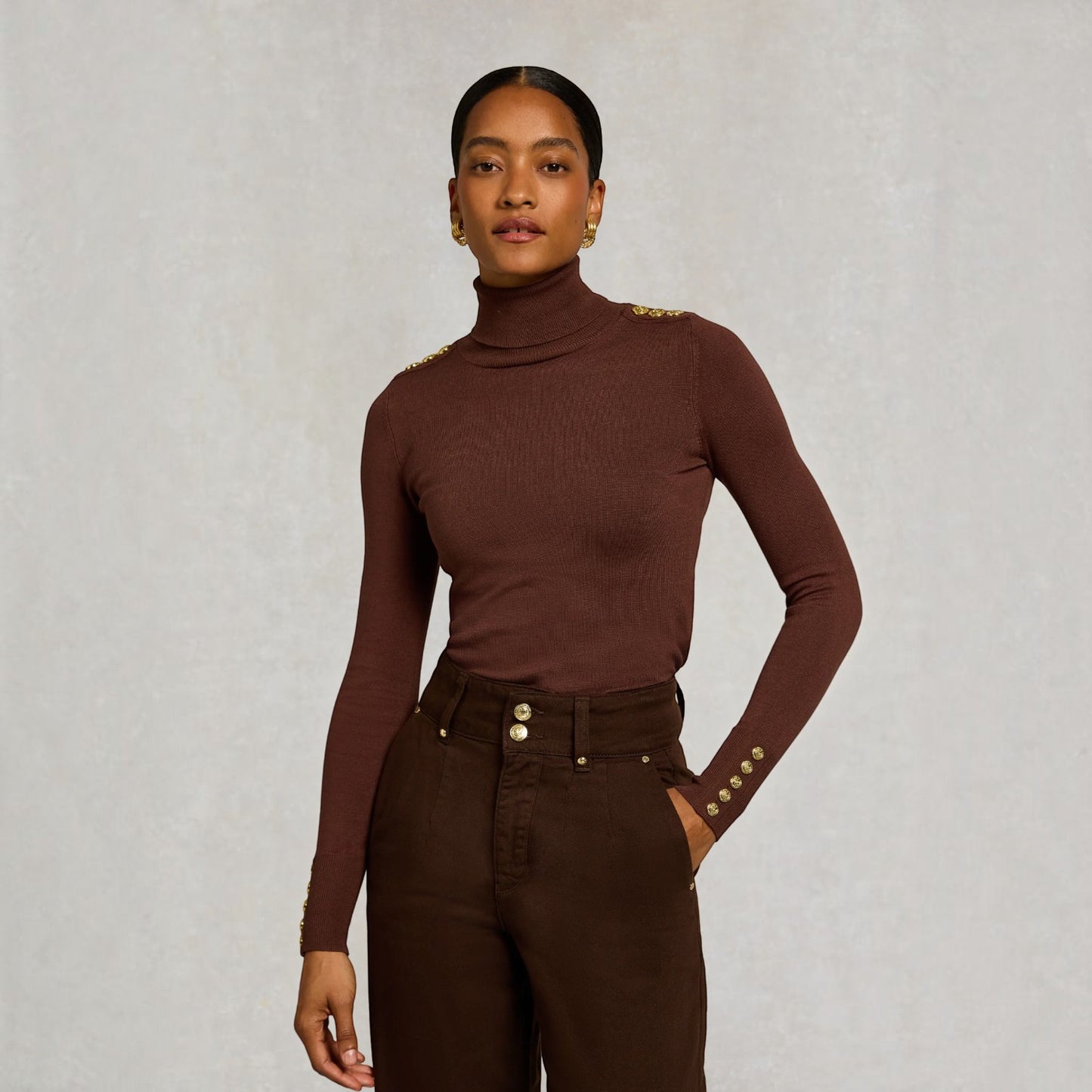 A model wearing a ribbed brown turtleneck top with gold button details on the shoulders and cuffs, paired with dark brown trousers, against a textured light gray background.