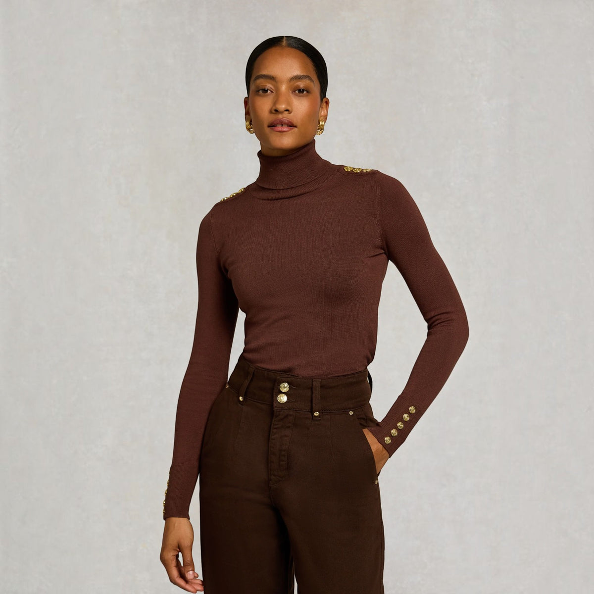 A model wearing a ribbed brown turtleneck top with gold button details on the shoulders and cuffs, paired with dark brown trousers, against a textured light gray background.