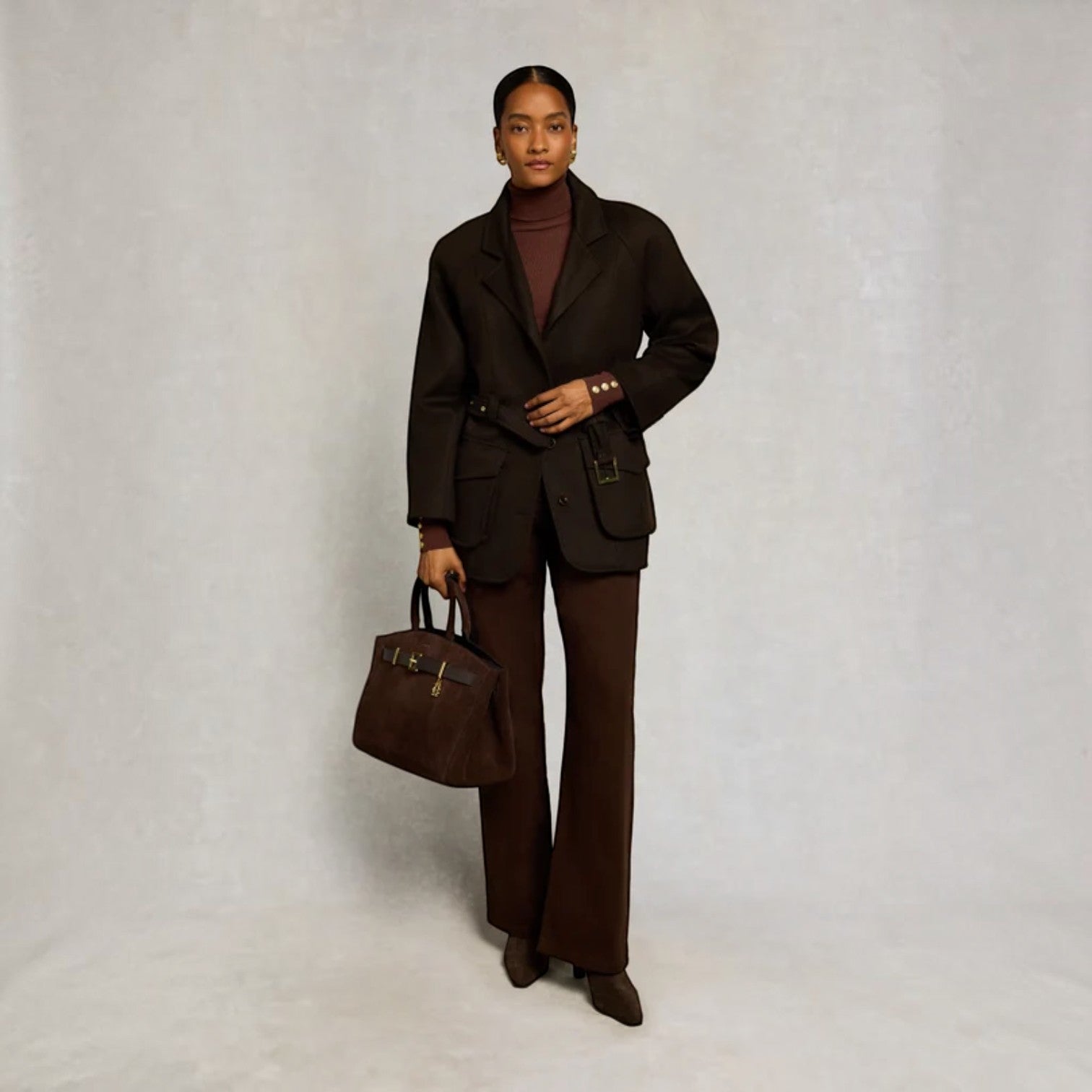 A model stands against a textured light gray studio backdrop wearing a dark brown tailored suit with a matching blazer, wide-leg trousers, and a dark brown turtleneck. She holds a dark brown structured handbag with gold-tone hardware.