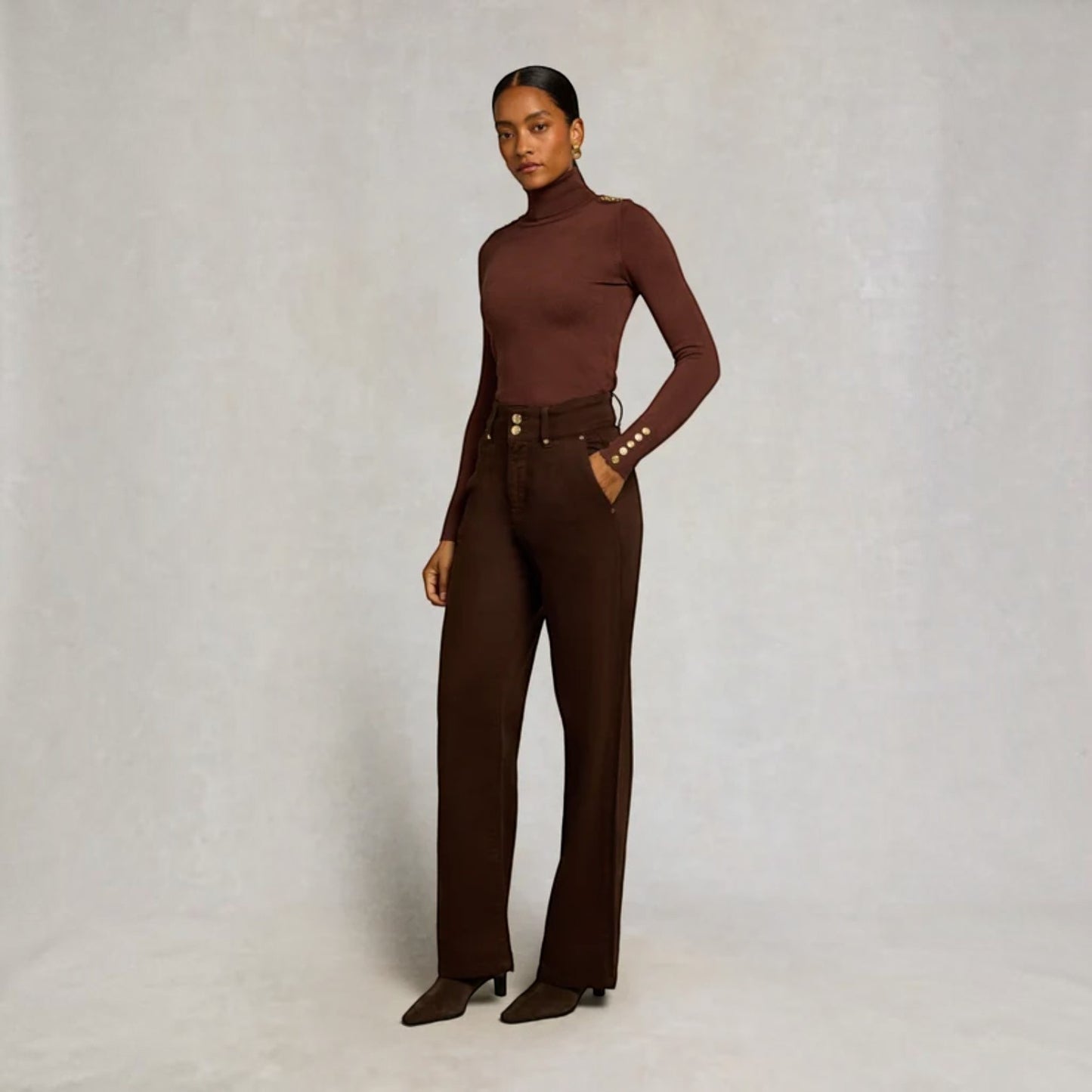 A model stands against a textured light gray studio backdrop wearing a fitted long-sleeve turtleneck top and matching high-waisted wide-leg trousers in a deep chocolate brown, styled with a matching belt and brown pointed-toe heels.