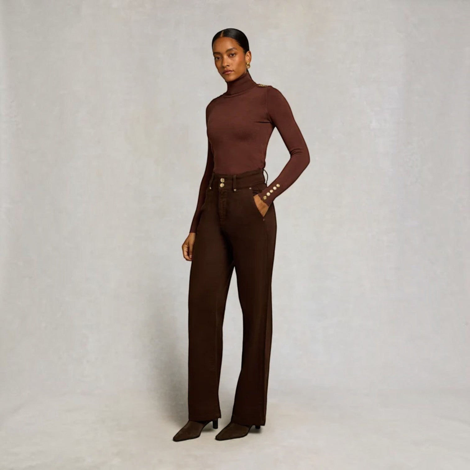 A model stands against a textured light gray studio backdrop wearing a fitted long-sleeve turtleneck top and matching high-waisted wide-leg trousers in a deep chocolate brown, styled with a matching belt and brown pointed-toe heels.