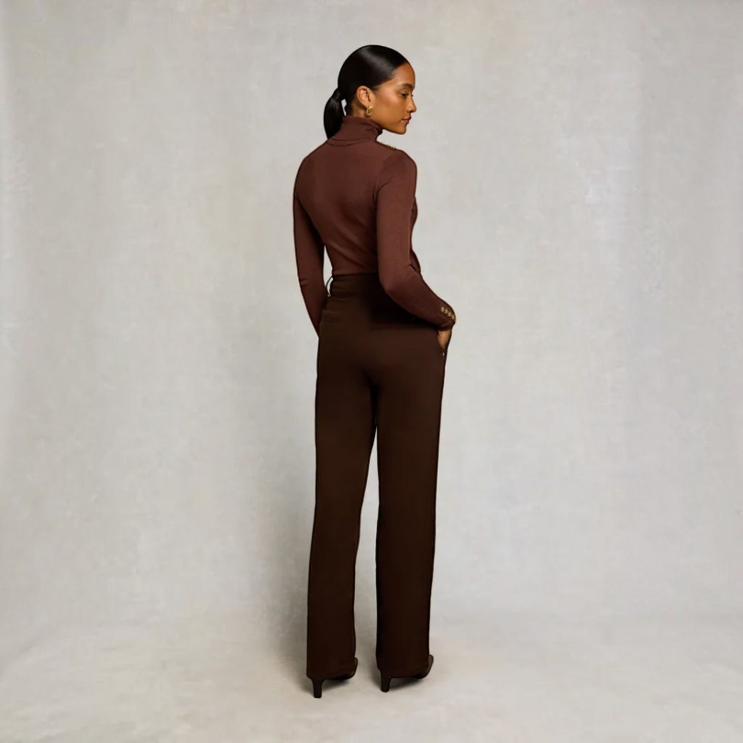 A woman stands with her back to the camera wearing a matching dark brown mock-neck long-sleeve top and wide-leg trousers set, photographed against a neutral textured studio backdrop.