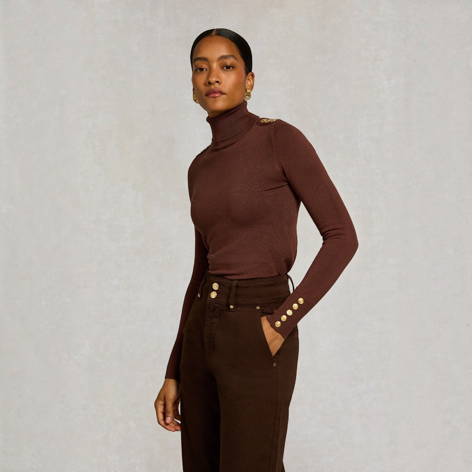 A model wearing a fitted, long-sleeve brown mock neck top with decorative gold buttons on the shoulders and cuffs, paired with dark brown trousers, set against a textured light gray background.