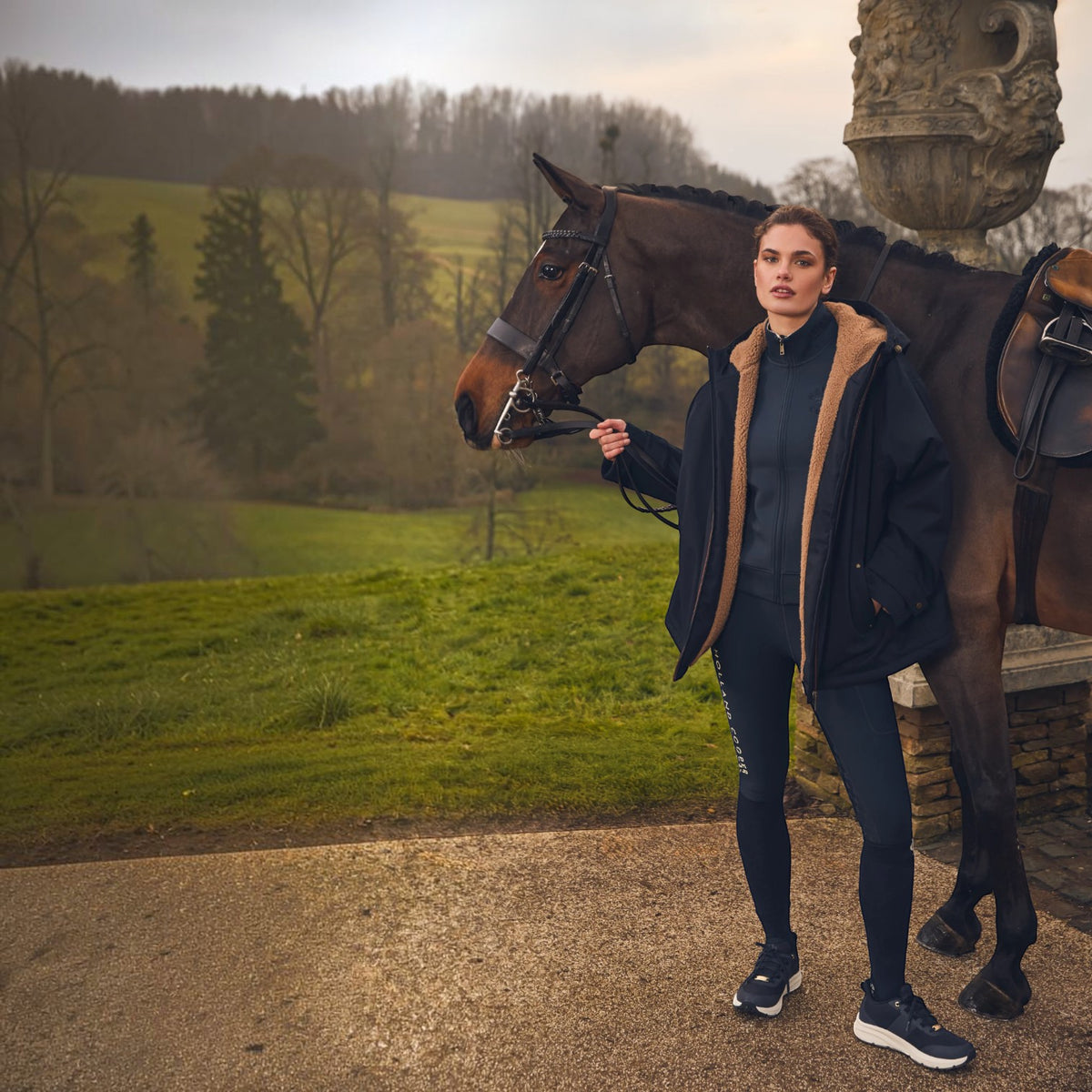 A woman stands outdoors beside a dark brown horse on a stone path, wearing a navy zip-front jacket with a tan fleece collar, a matching navy top and leggings, and dark sneakers. The background shows a grassy field, trees, and a stone architectural element.