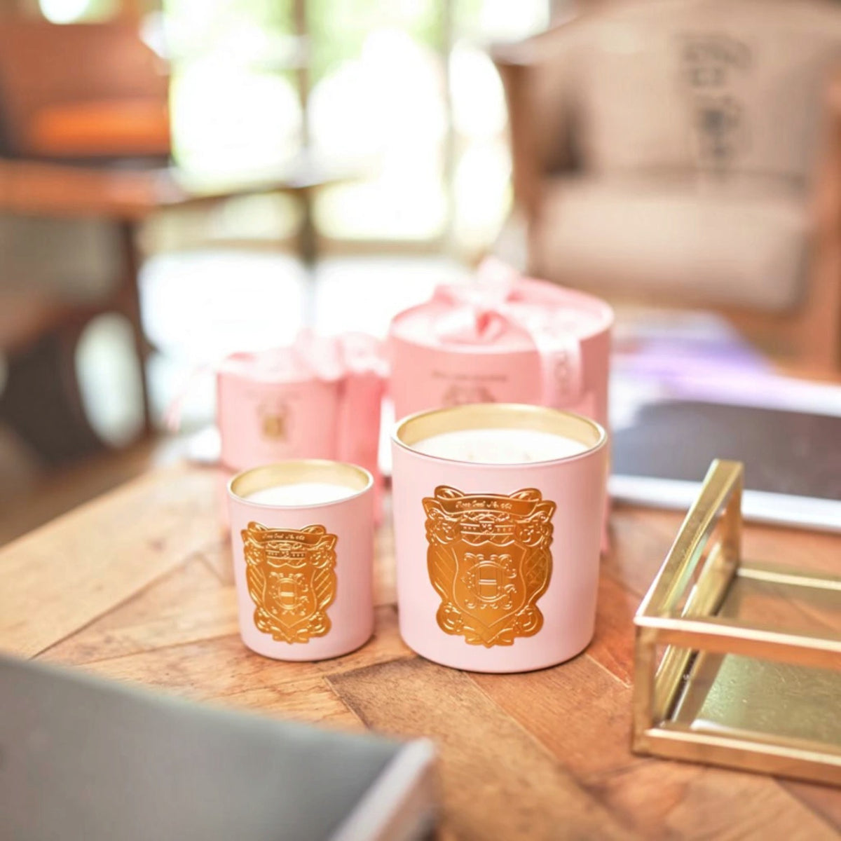 Two pink metal candle jars with gold crest designs on the front, displayed on a wooden table with matching pink gift boxes in a softly blurred indoor setting.