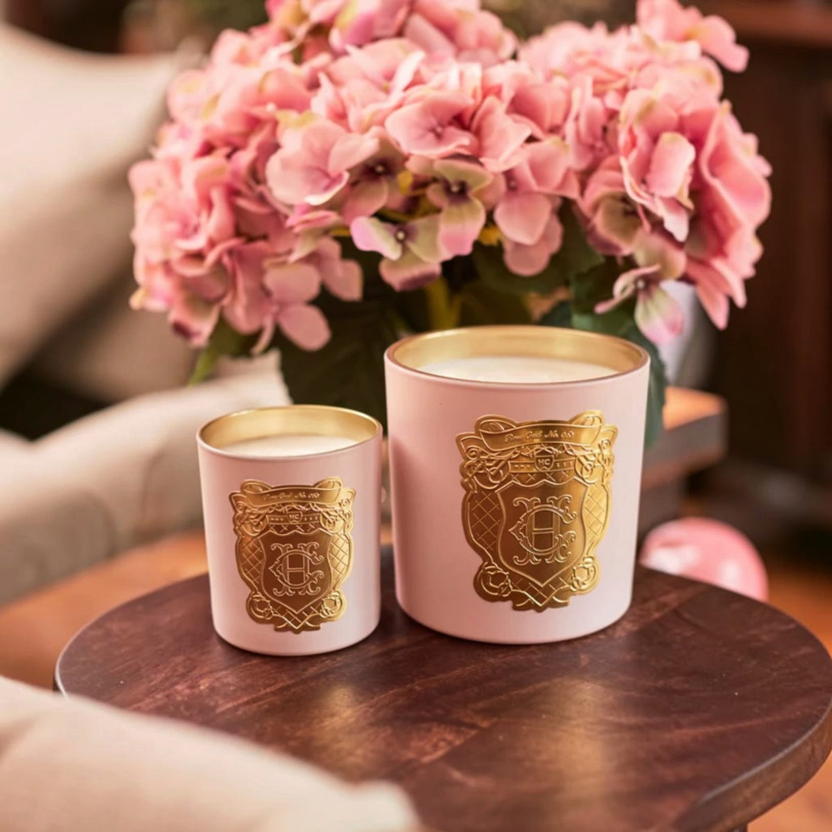 Two pink wax candles with gold crest labels sit on a dark wooden table, with a blurred background of pink flowers and a sofa.