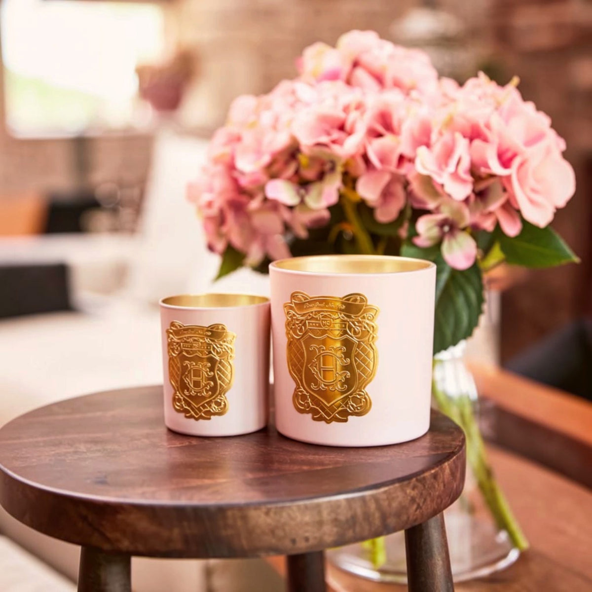 Two pink wax candles with gold foil crest designs sit on a round wooden table, with a vase of pink flowers blurred in the background.