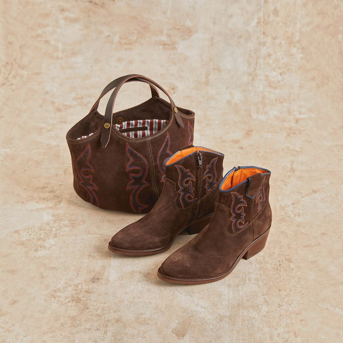A matching brown suede western boot and tote bag set on a textured beige stone background. The boots feature dark brown western embroidery, a zippered ankle opening, and bright orange lining, while the matching tote bag has a plaid lining and a single top handle.