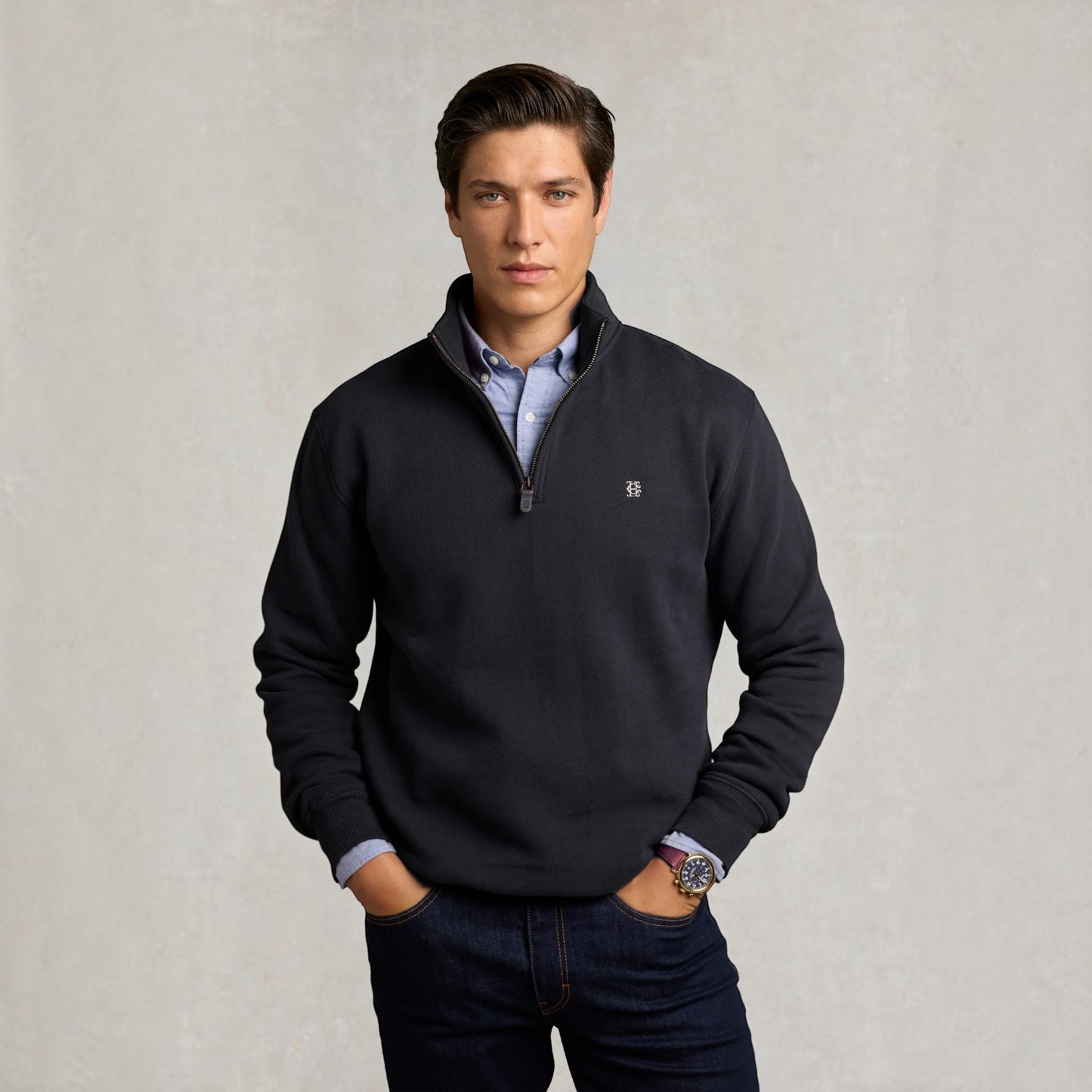 A man wearing a dark navy quarter-zip pullover sweater over a light blue collared shirt and dark jeans, standing against a textured light gray background.