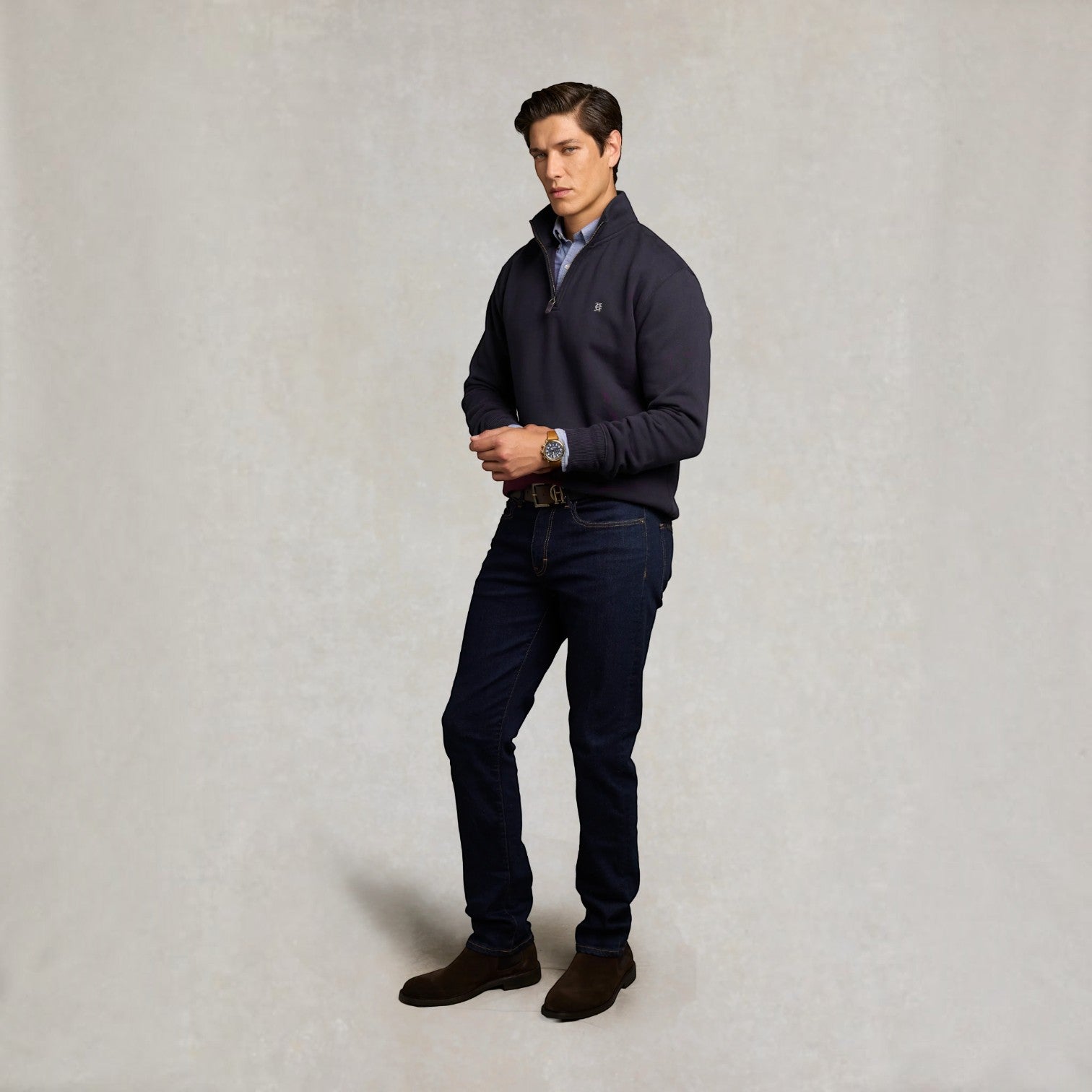 A man modeling a dark navy quarter-zip pullover sweater, paired with dark blue jeans, a light blue patterned shirt, and brown suede boots, standing against a textured light gray studio backdrop.