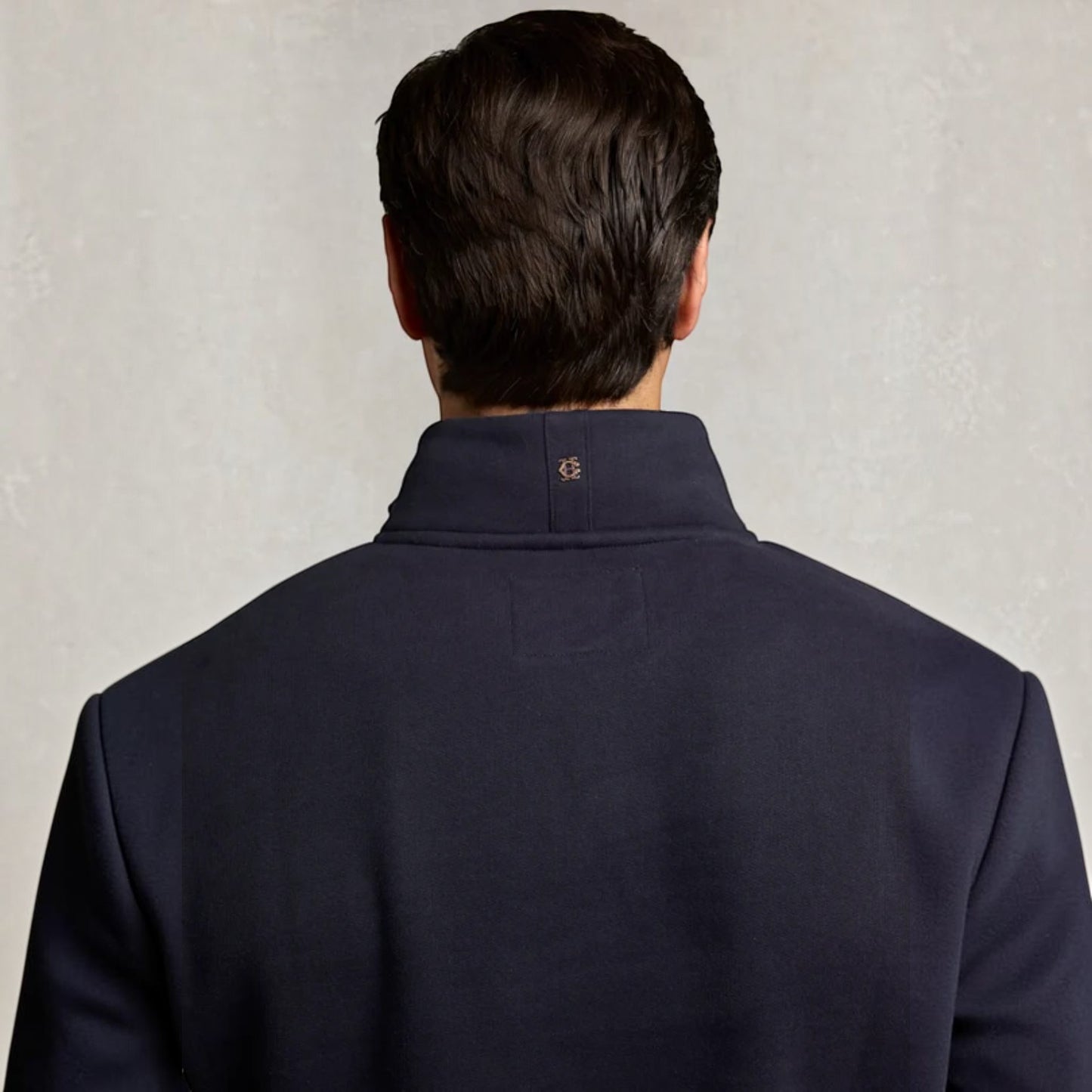 Rear view of a man wearing a navy blue high-collar pullover jacket against a textured light gray background.
