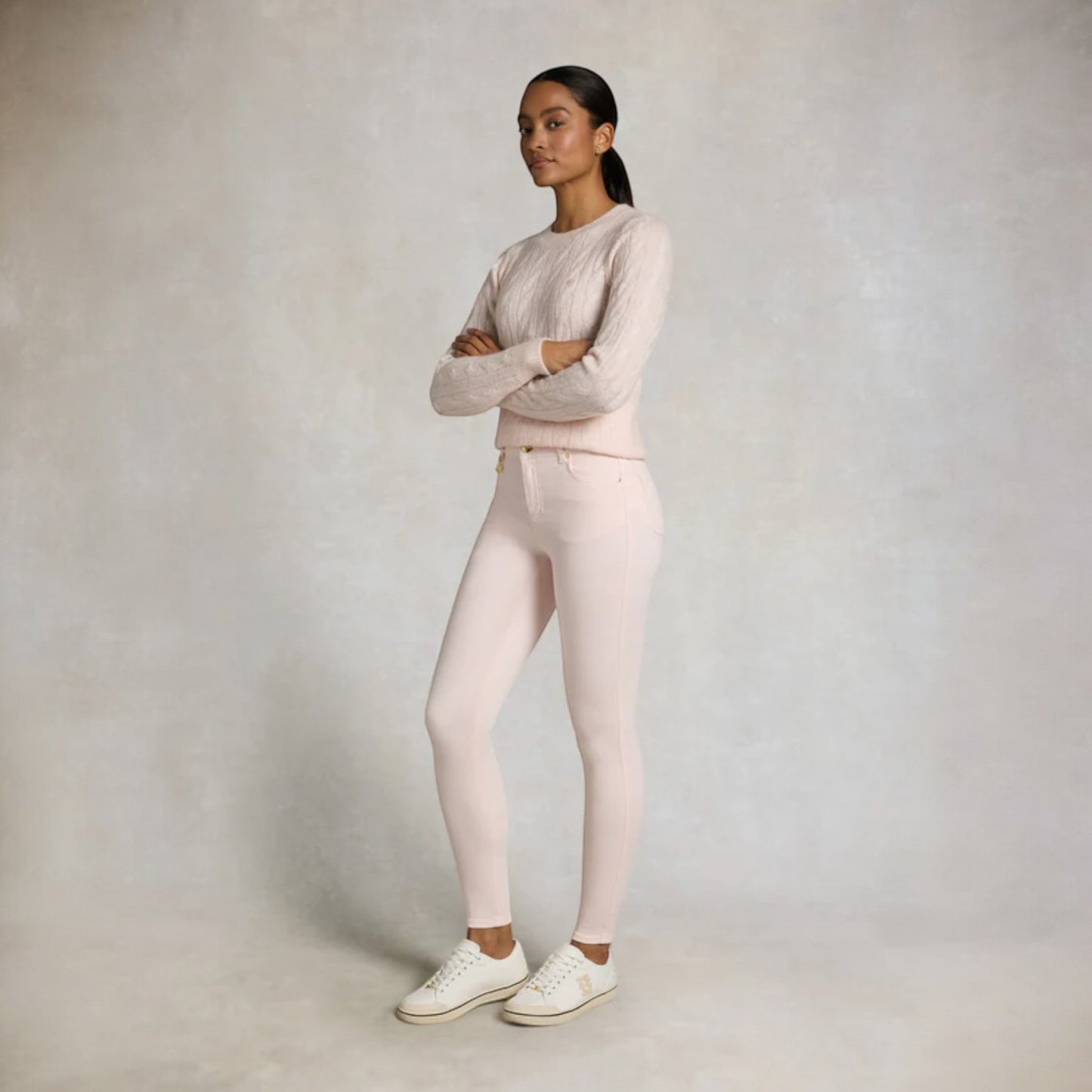 Woman wearing a light pink sweater and pants with white shoes on a plain background