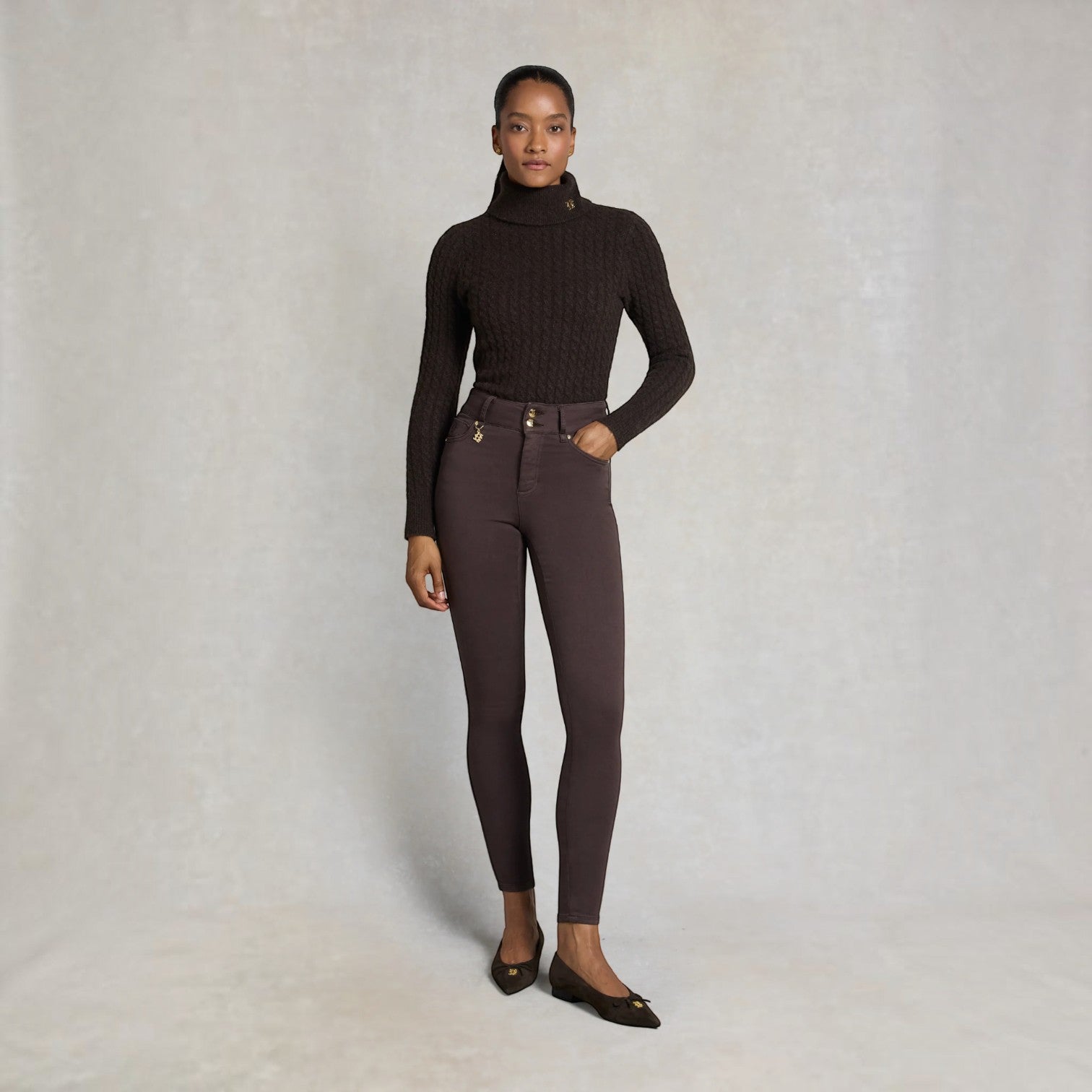 A model stands against a textured light gray studio backdrop wearing a dark brown cable-knit turtleneck sweater, dark brown skinny pants with gold-tone hardware, and dark brown flats.