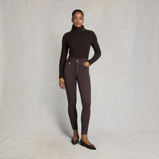 A model stands against a textured light gray studio backdrop wearing a dark brown cable-knit turtleneck sweater, dark brown skinny pants with gold-tone hardware, and dark brown flats.