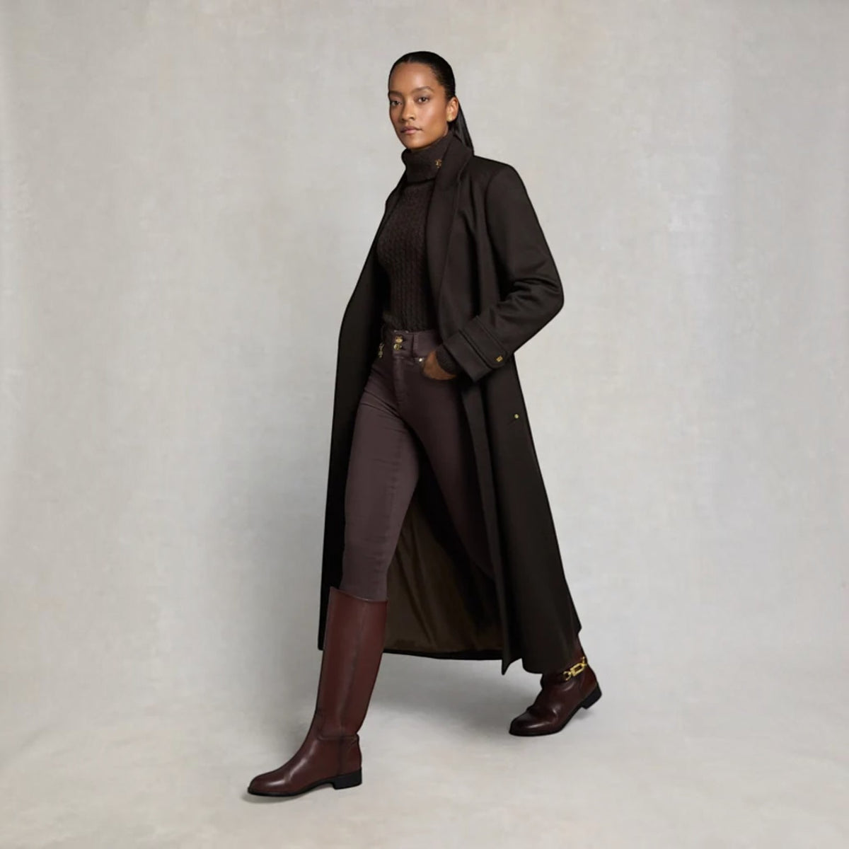 A model wearing a long dark brown overcoat over a dark brown turtleneck, matching brown pants, and dark brown knee-high boots, standing against a neutral light gray studio background.