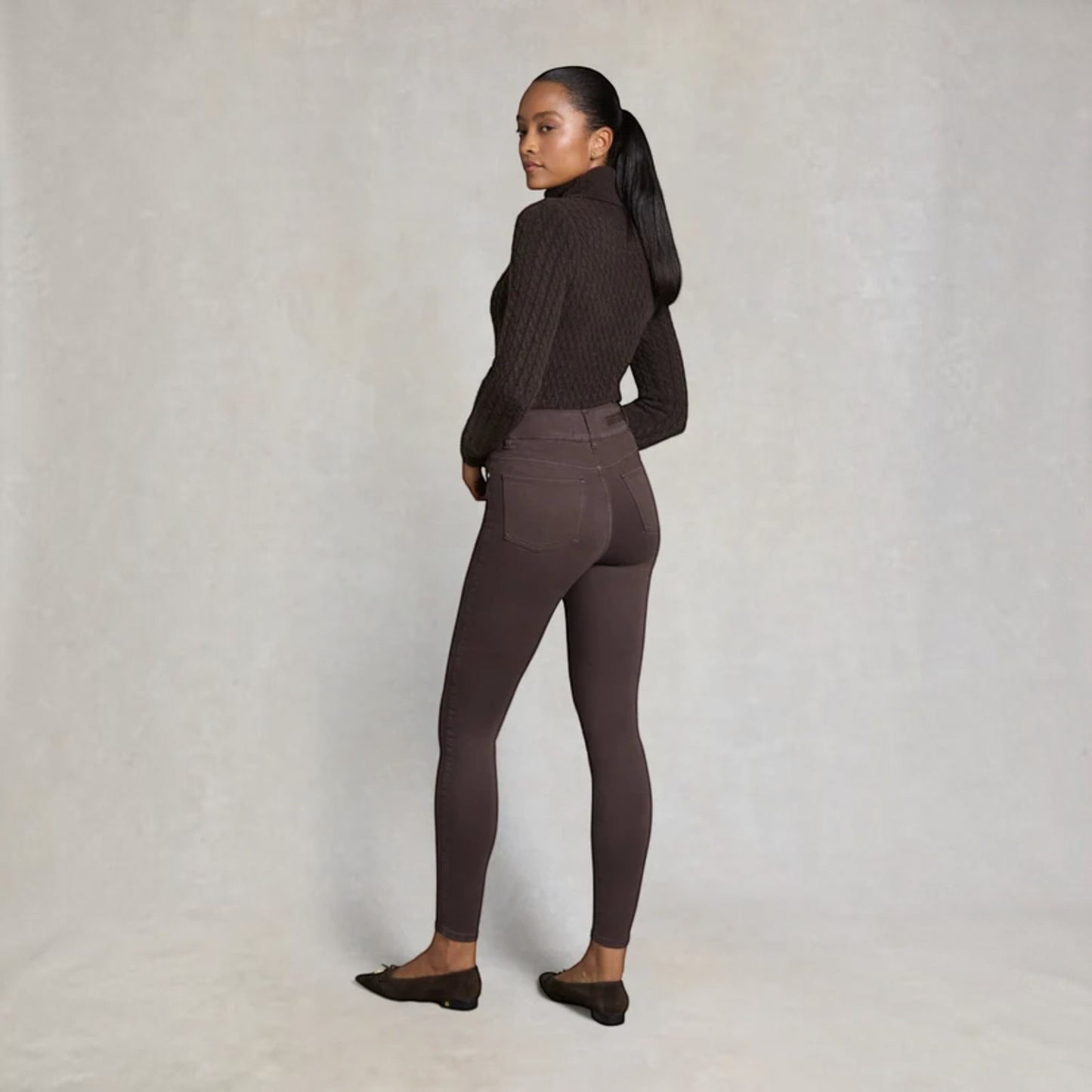 A woman stands in profile wearing a dark brown ribbed turtleneck sweater and matching dark brown leggings, paired with dark flats, against a neutral textured studio backdrop.