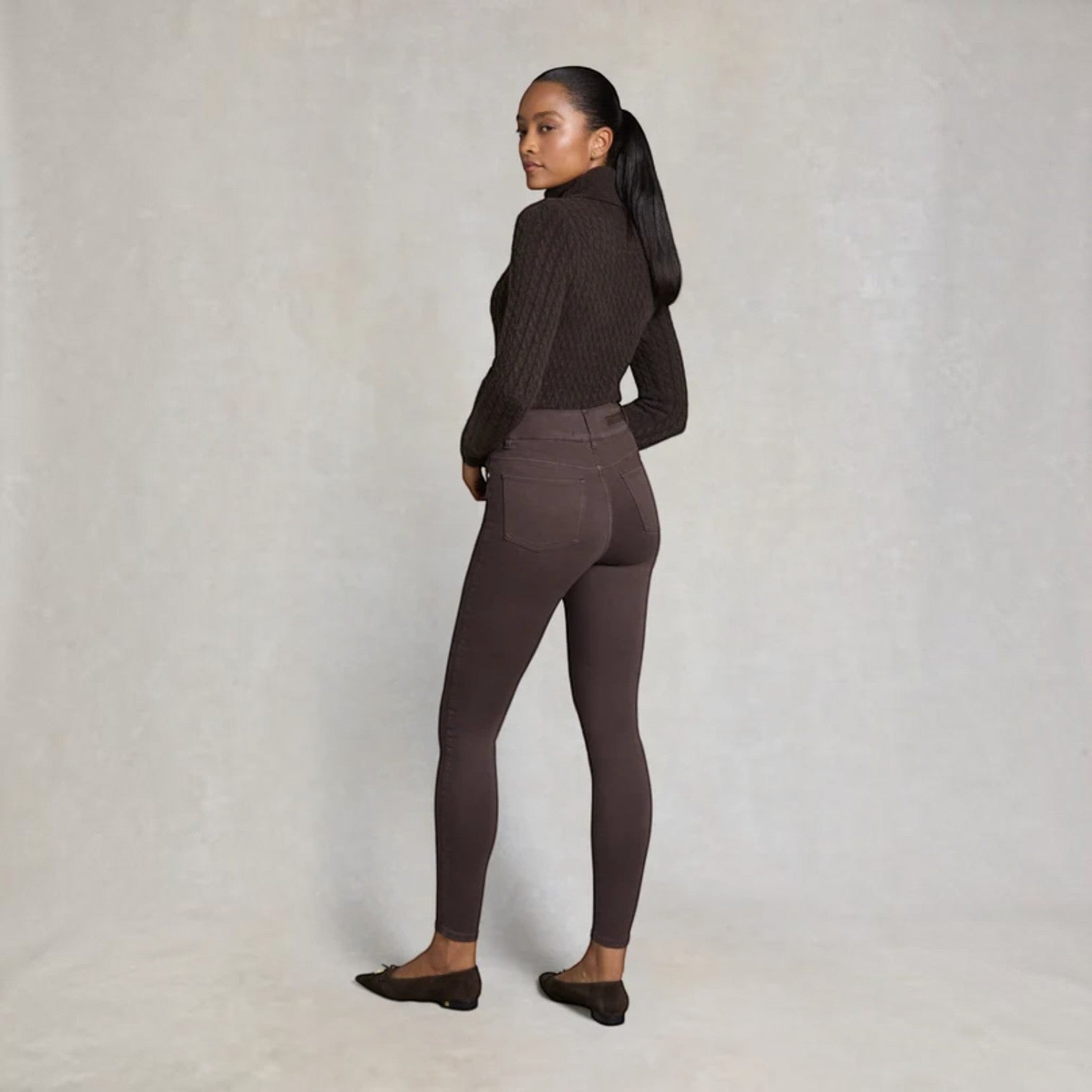 A woman stands in profile wearing a dark brown ribbed turtleneck sweater and matching dark brown leggings, paired with dark flats, against a neutral textured studio backdrop.