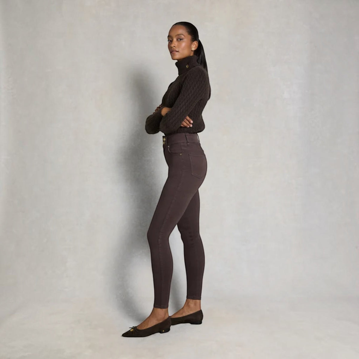 A woman stands in profile wearing dark brown skinny jeans, a dark brown cable-knit mock-neck top, and dark brown flats against a textured light gray studio backdrop.