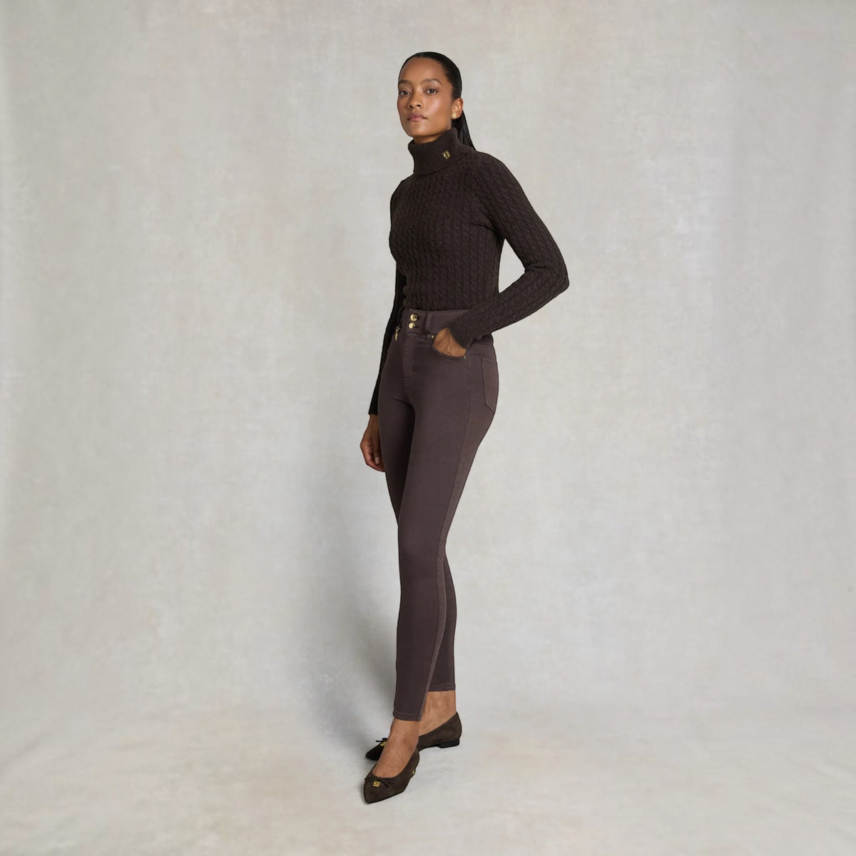 A woman modeling a dark brown ribbed mock-neck long-sleeve top and matching dark brown fitted pants against a neutral textured studio backdrop.