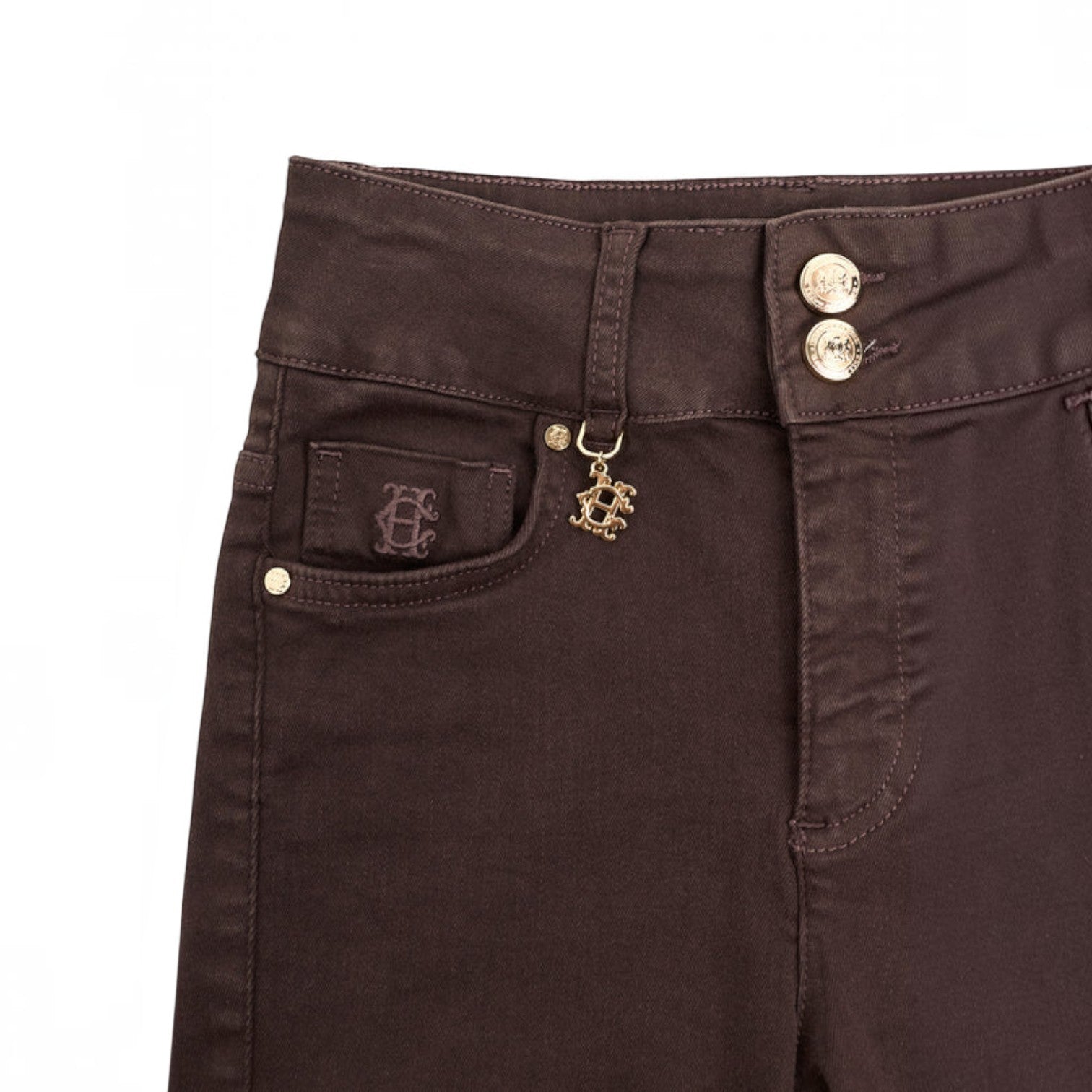 Close-up of dark brown denim jeans showing the front pocket with a monogram patch, a decorative gold-tone monogram charm, and double gold-tone buttons on the waistband.