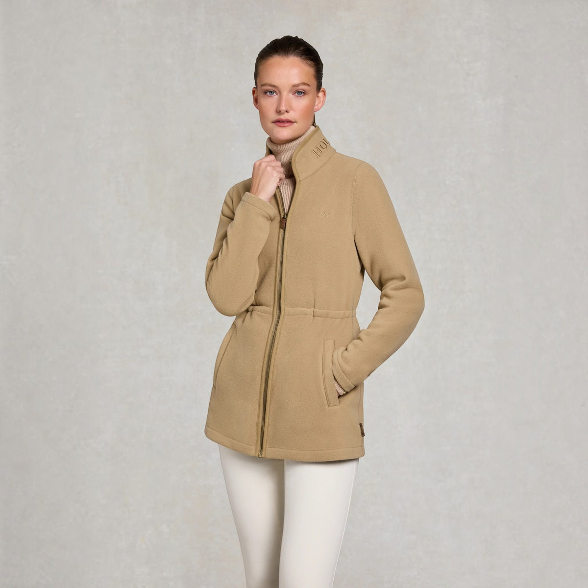A woman modeling a beige fleece jacket with a high collar, front zipper, and drawstring waist, paired with white pants, against a textured light gray studio background.