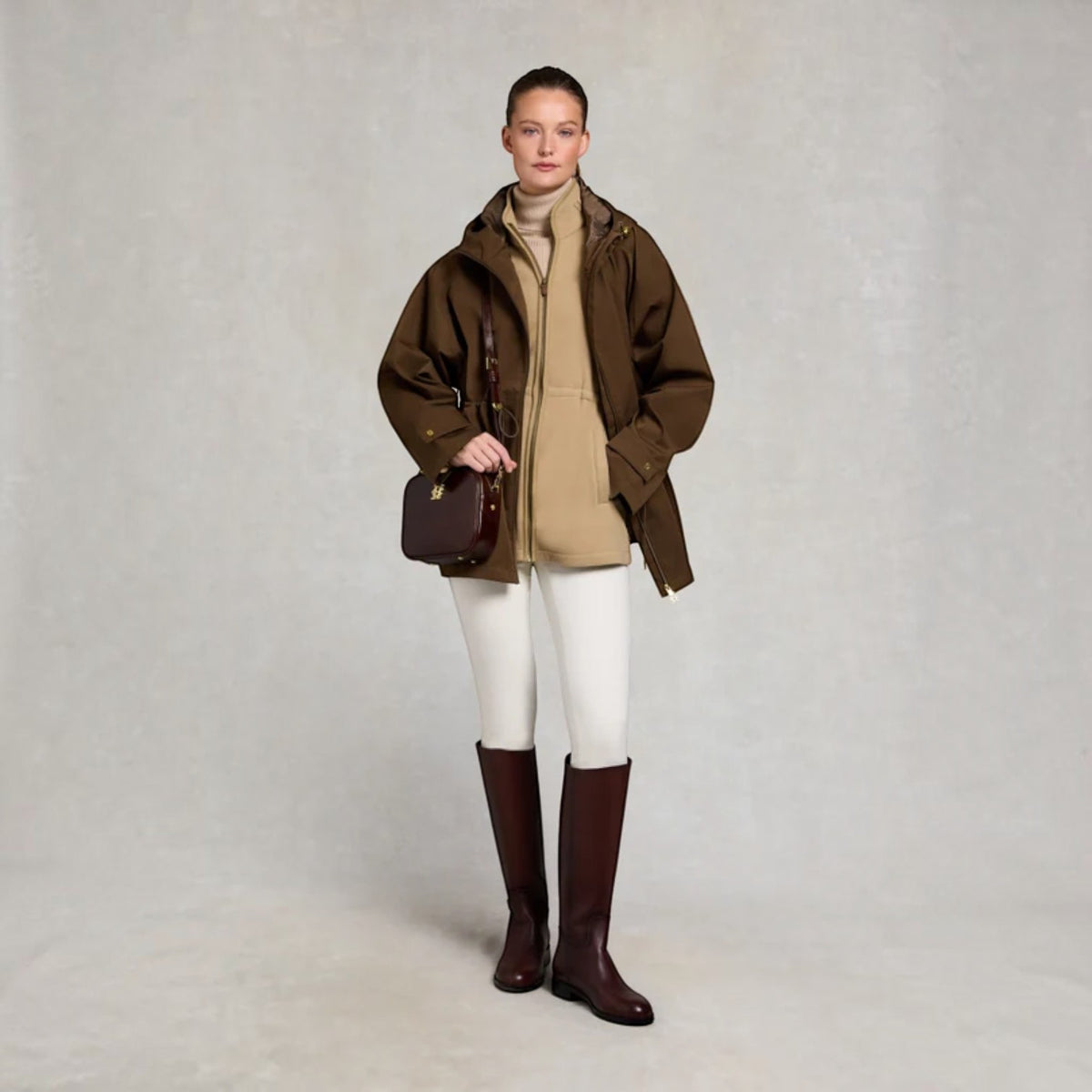 A model stands against a neutral light gray studio background wearing an olive brown hooded jacket over a beige turtleneck, white riding breeches, and dark brown knee-high boots, while holding a dark brown leather shoulder bag.