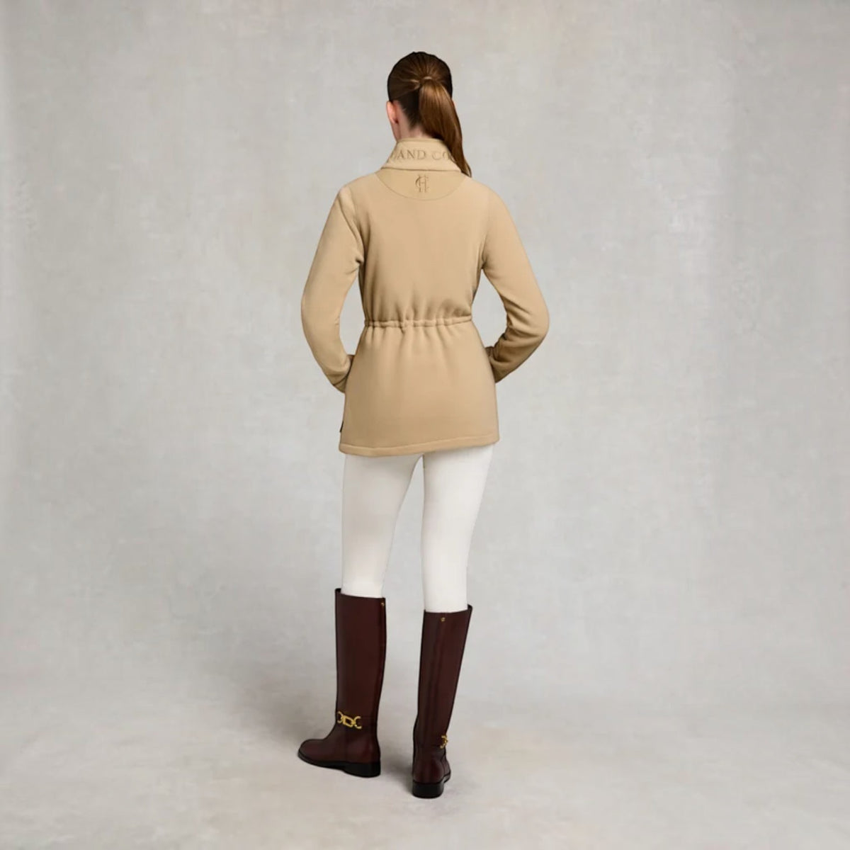 A woman stands with her back to the camera wearing a beige equestrian jacket with a drawstring waist, white breeches, and dark brown riding boots against a neutral gray studio backdrop.