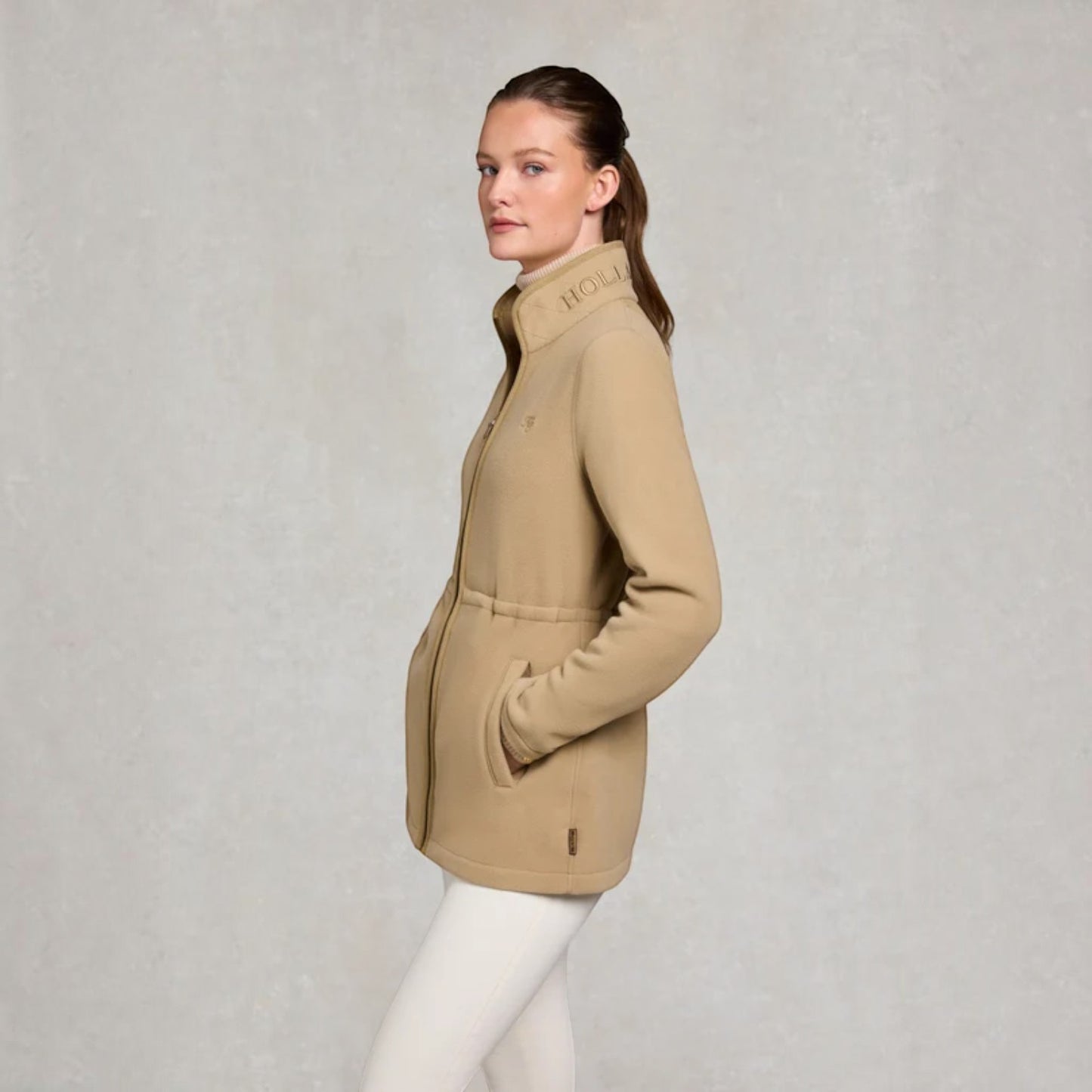 A woman wearing a beige mid-length zip-up jacket with a high collar and white pants, standing against a textured light gray studio background. The brand name Holland cooper is visible on the collar.