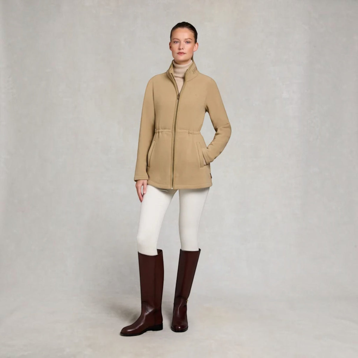 A woman stands against a light gray textured studio backdrop wearing a camel-colored zip-front jacket with a stand collar, white fitted pants, and dark brown knee-high boots.