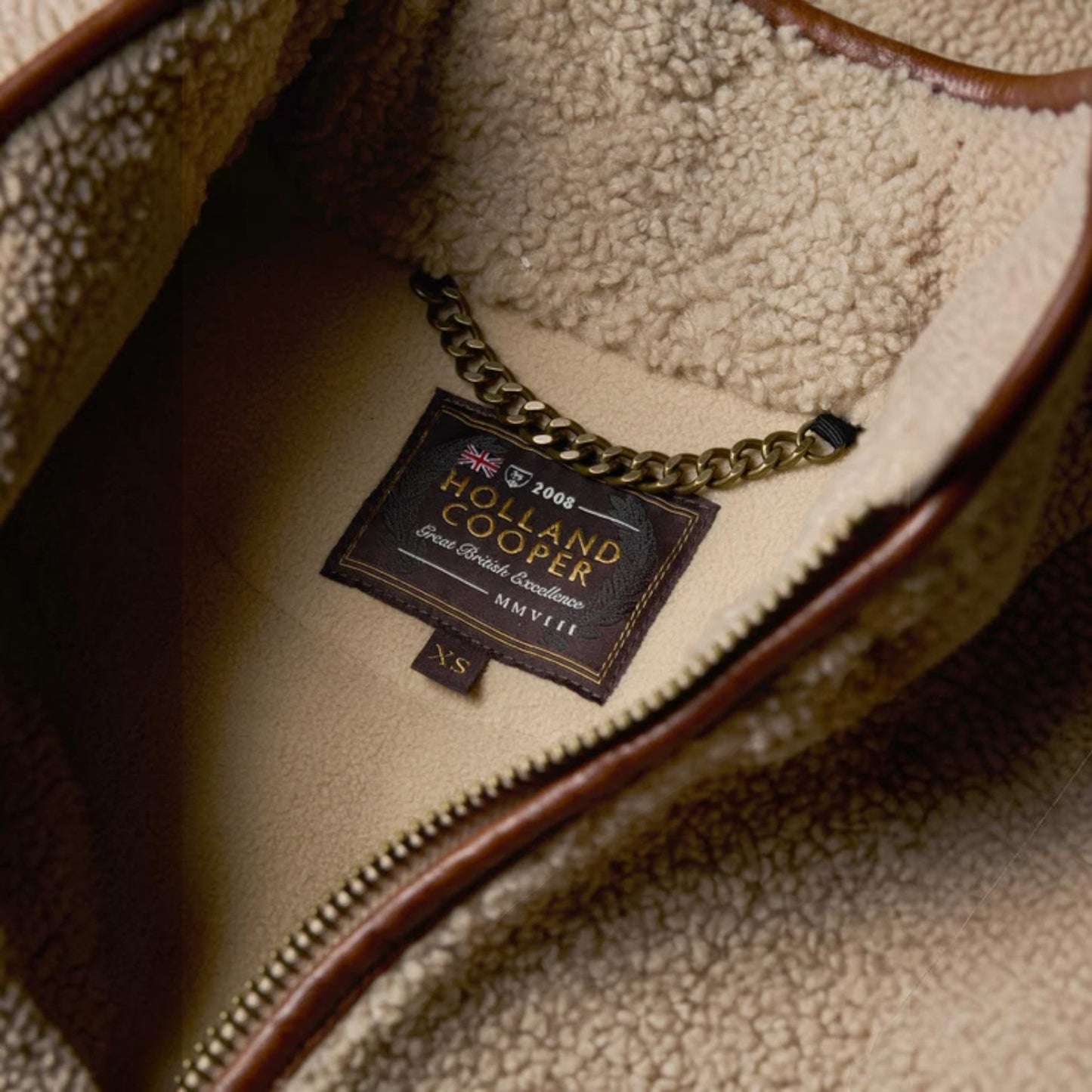 Close-up of the interior of a brown and beige fleece bag, showing a sewn-in brand label that reads Holland Cooper with a gold chain attached.