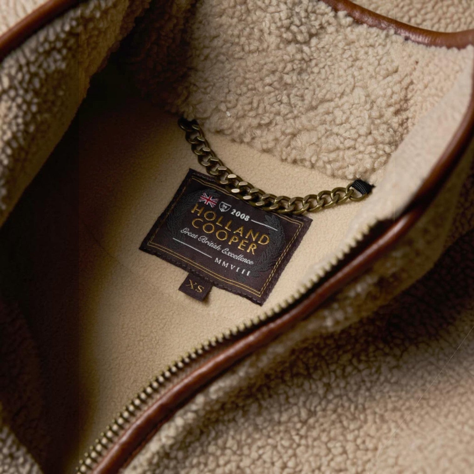 Close-up of the interior of a brown and beige fleece bag, showing a sewn-in brand label that reads Holland Cooper with a gold chain attached.