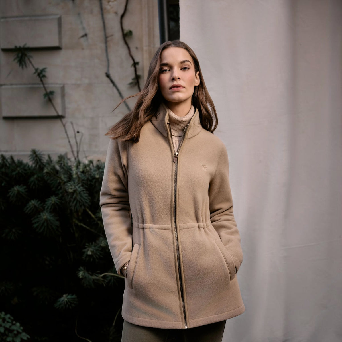 A woman wearing a beige zip-up coat over a matching beige turtleneck sweater, standing outdoors against a light gray wall and dark green foliage.