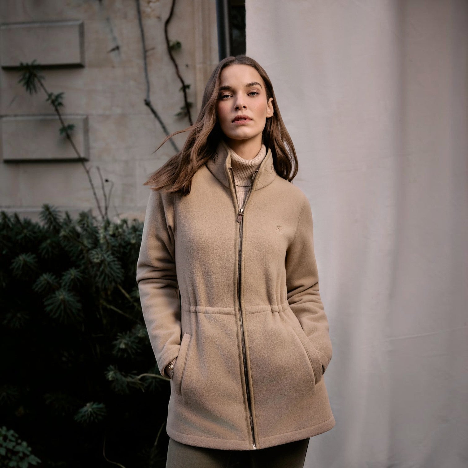 A woman wearing a beige zip-up coat over a matching beige turtleneck sweater, standing outdoors against a light gray wall and dark green foliage.